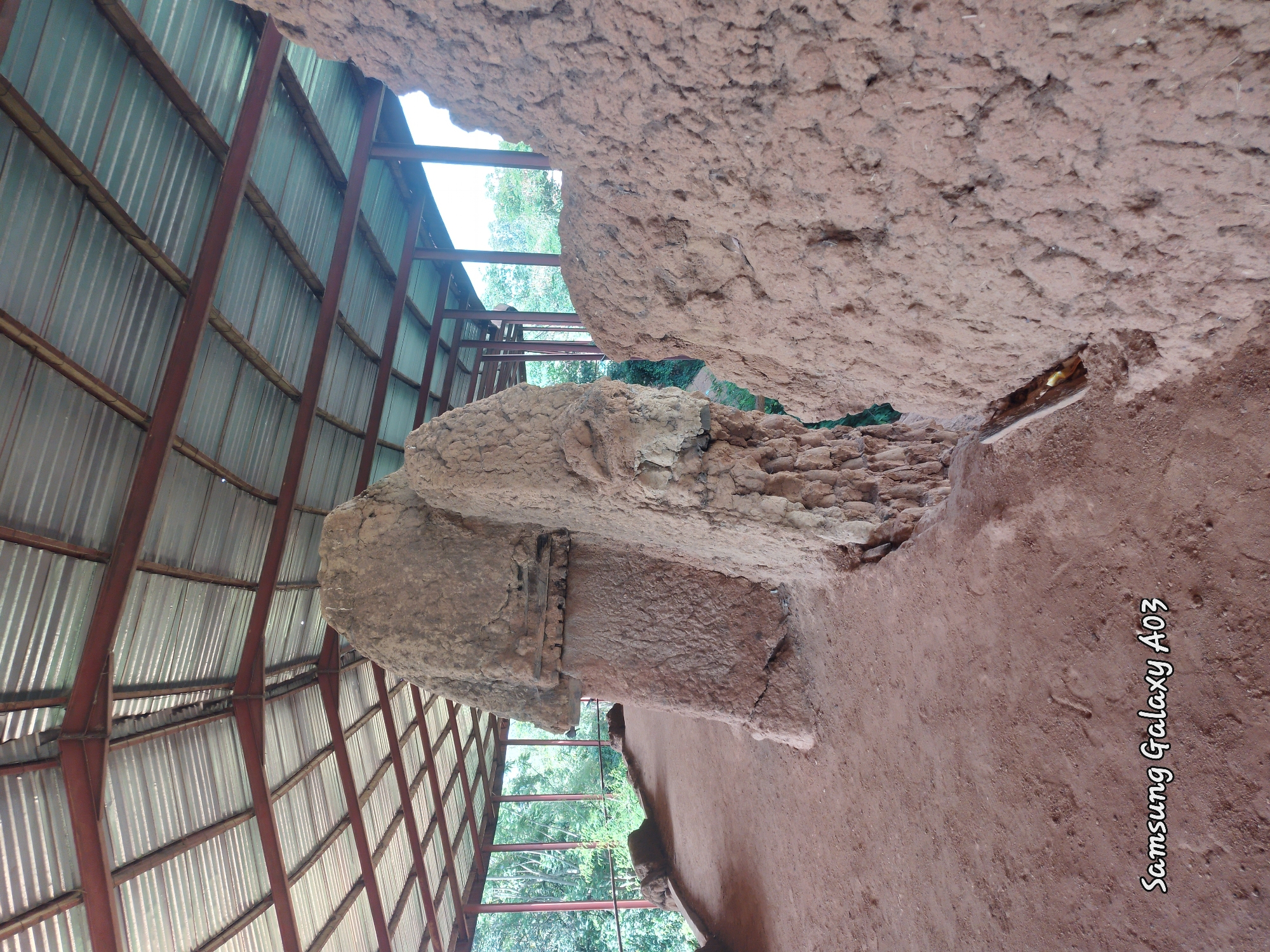 The image shows a view of the ancient stone sculptures of the Tangle in the city of Jos, Nigeria, under a protective corrugated iron roof supported by metal beams. The sculptures are made of reddish-brown clay-like material, with rough, textured surfaces. Some of the stones are large and imposing, with visible layers and cracks. The ground in front of the sculptures is also made of reddish-brown earth, with some footprints visible. In the background, lush green trees and vegetation can be seen through the open sides of the shelter, suggesting an outdoor environment. The sky is overcast. A watermark in the bottom left corner reads "Samsung Galaxy A03".