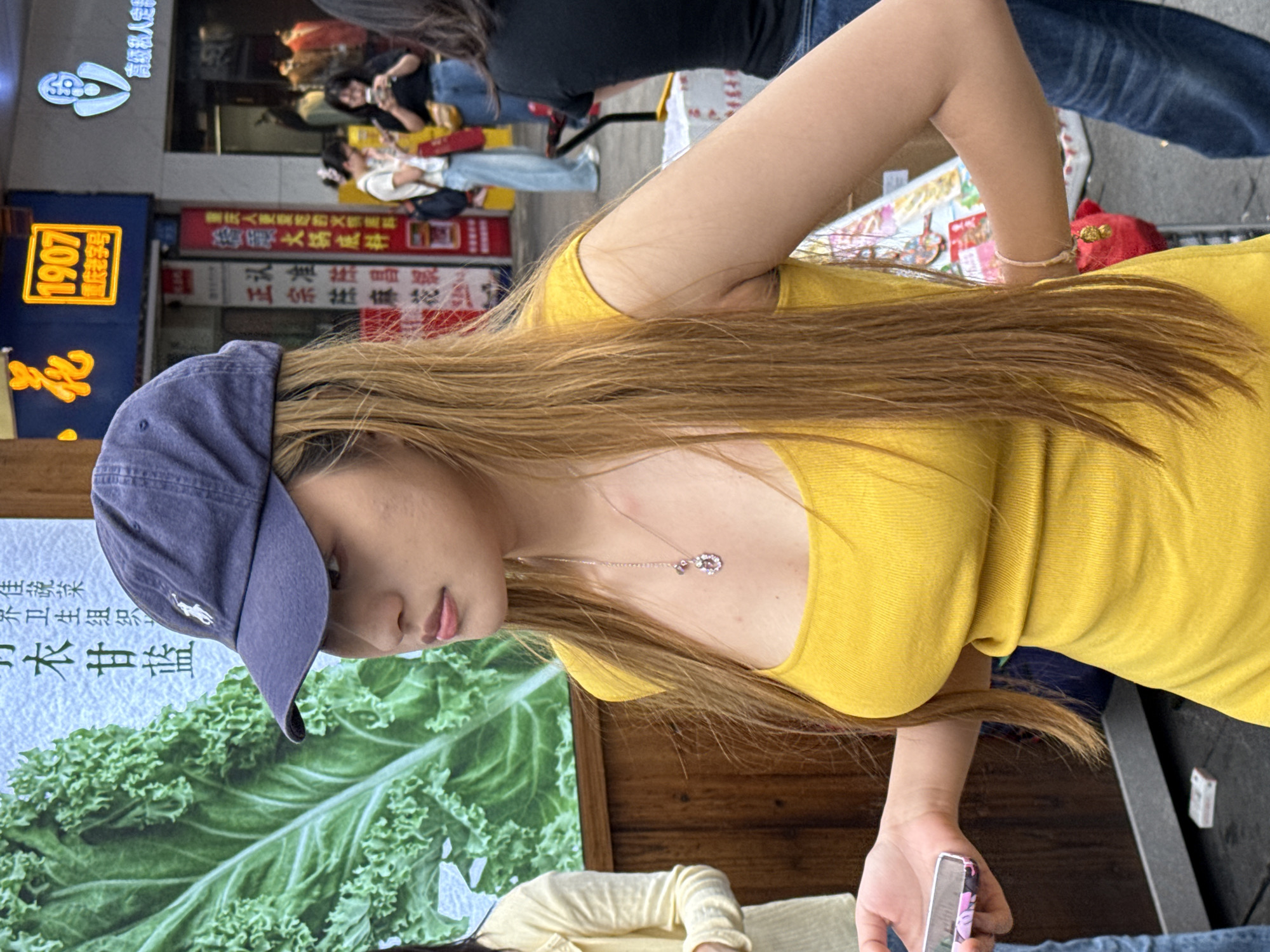 A young woman with long blonde hair, wearing a blue baseball cap and a yellow top, is seen outdoors. She is looking down and holding a phone in her right hand. In the background, there are various signs written in Chinese. One sign reads "1907 Chongqing Old Brand" and includes the text "Chongqing People Like to Eat Fire Material". The woman is standing next to a poster featuring an image of kale. Other people are visible in the background. The scene appears to be in a bustling urban environment, likely within the city of Liangjiang, China.