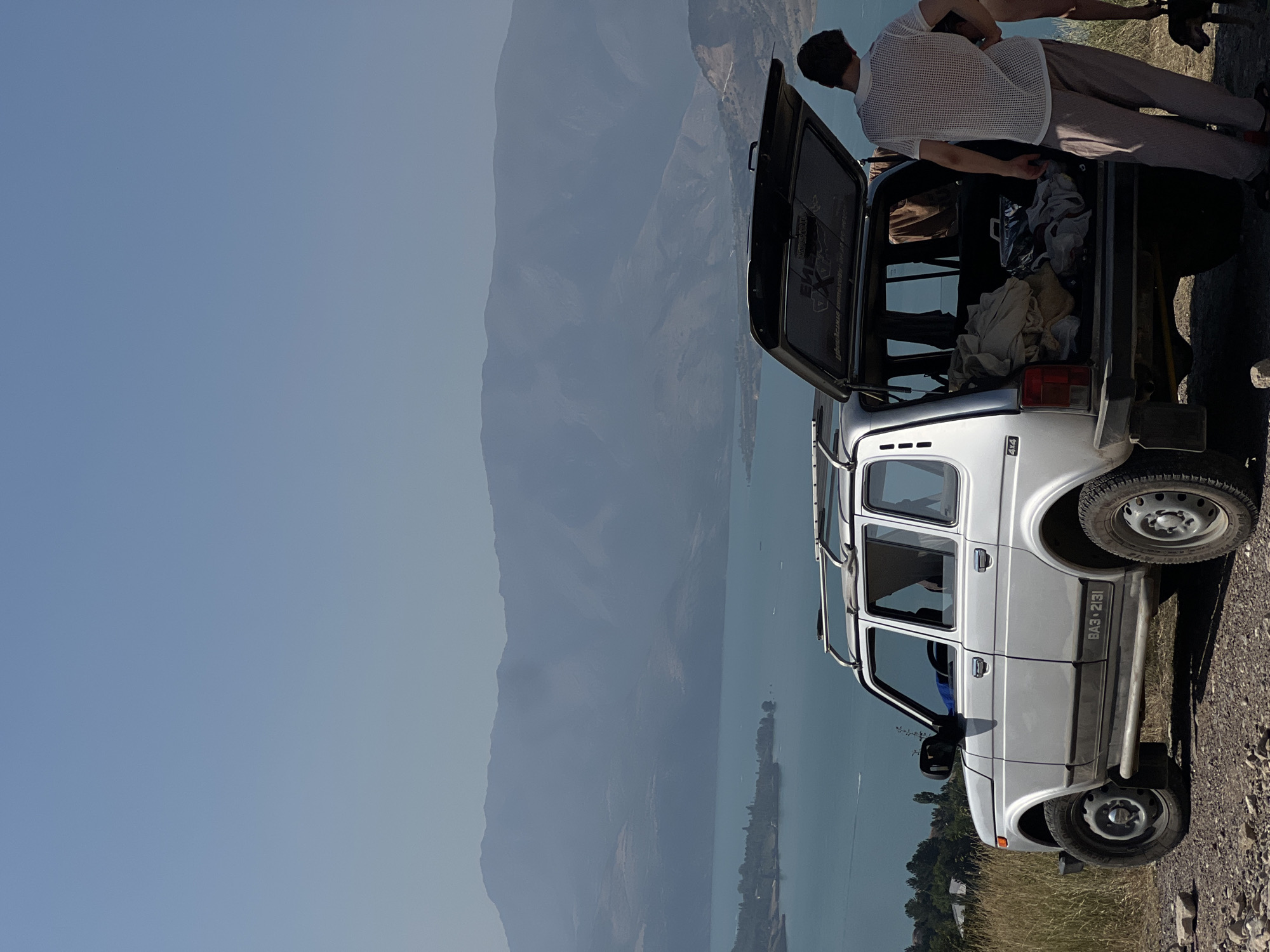 A silver Niva vehicle is parked on a gravelly embankment overlooking a large, turquoise body of water, likely a reservoir. The vehicle's rear hatch is open, revealing the interior filled with various items, including bags and folded clothing. A young man, wearing a white mesh t-shirt and striped pants, stands by the open hatch, facing away from the camera and looking towards the water. In the background, hazy mountains rise majestically, with a lighter blue sky above. The environment appears dry and arid, with sparse vegetation on the embankment. A small dog is visible near the man's feet. The scene suggests a stop during a road trip or an outdoor excursion. The location is described as the city of Yangikurgan, Uzbekistan.