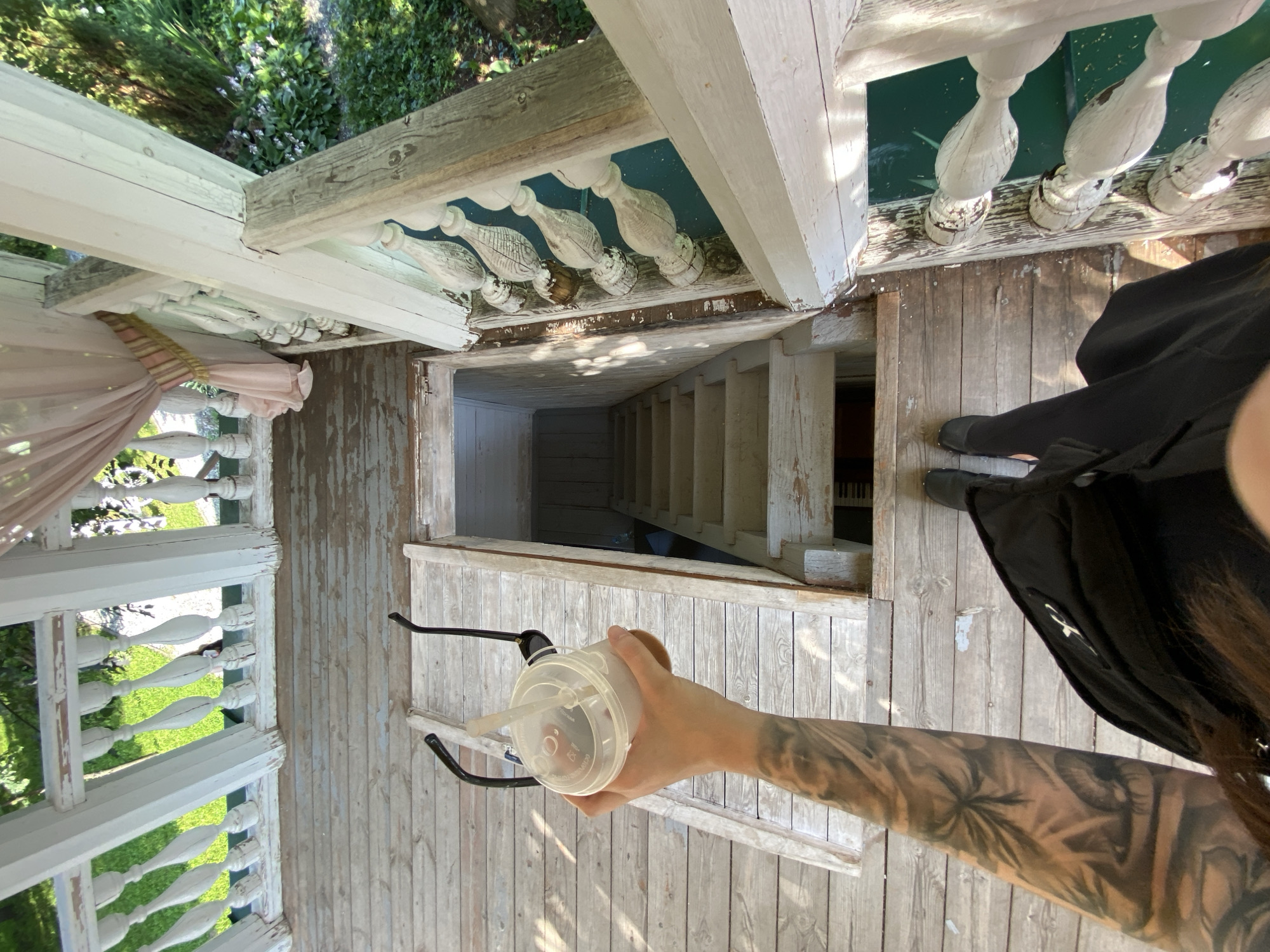 The image depicts a person's arm holding a plastic cup with a straw and sunglasses hooked onto it, standing on a wooden porch in Votkinsk, Russia. The porch has white balusters and a wooden railing. Below the porch, a set of stairs leads down into a dark opening. The person's legs, clad in black pants and shoes, are visible at the bottom of the frame. The environment suggests an outdoor setting with greenery visible in the background.