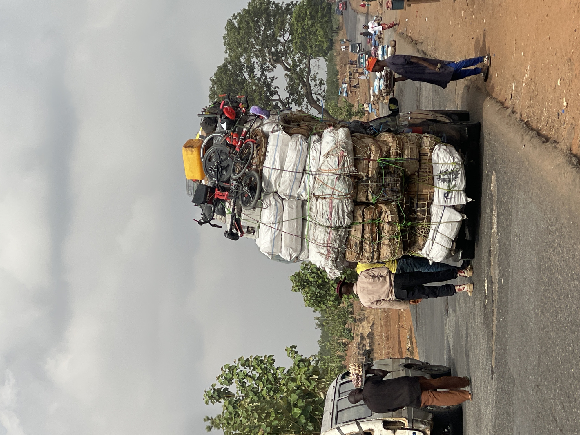 The image depicts a vehicle heavily overloaded with goods in Gidan Waya, Nigeria. The vehicle, a dark-colored sedan, is almost entirely obscured by a towering stack of goods including tightly bound packages, bicycles, a yellow canister and other items. Several people stand near the vehicle; One individual wearing a blue robe and orange cap stands near the car, and another two people, dressed in casual attire, are standing next to the car. Near the front of the car is a white van with one visible person. The road is paved and stretches into the distance, where other vehicles and people are visible. The background includes trees and a cloudy sky.