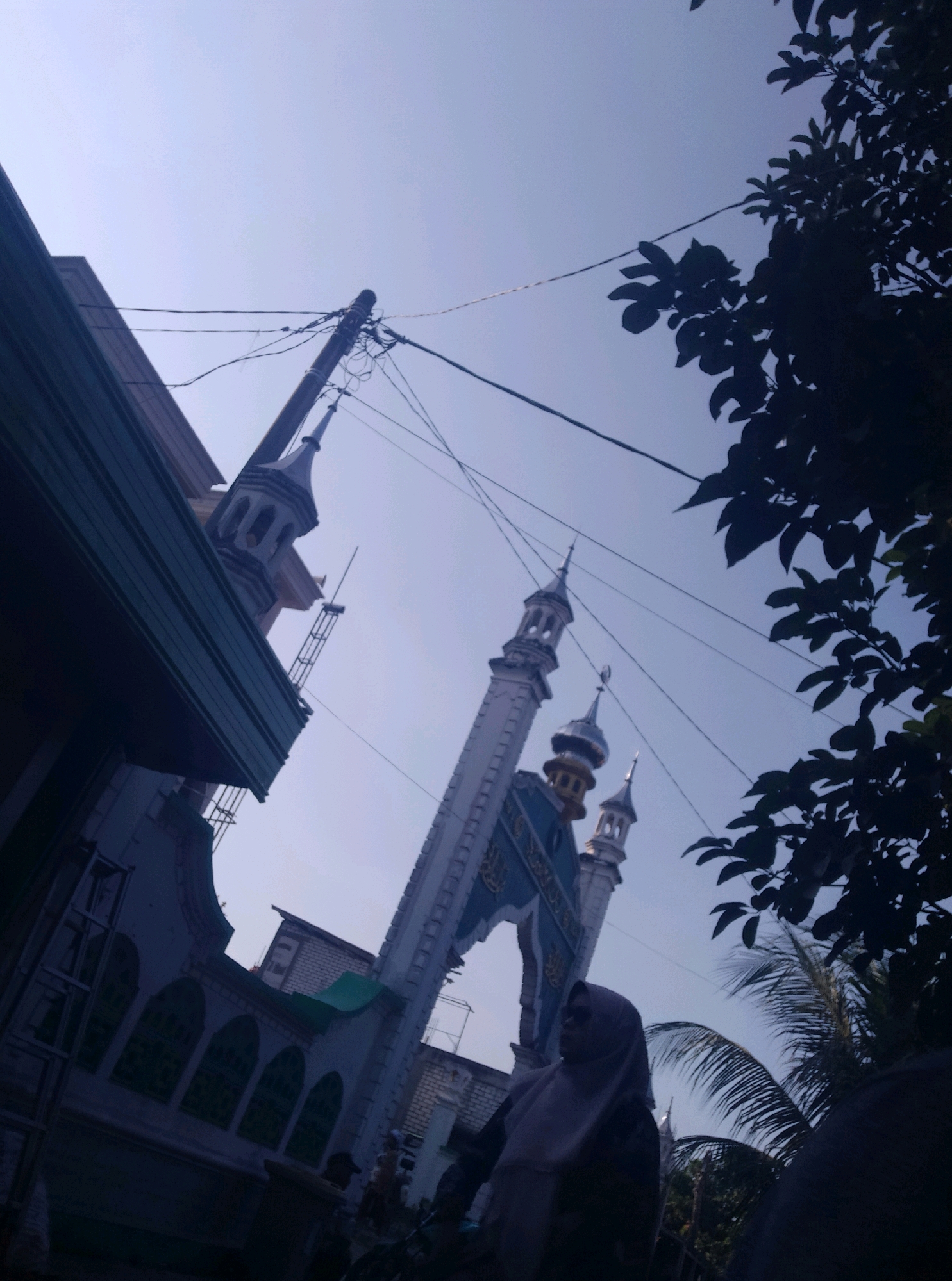 The image captures a view of a mosque in Wotan, Indonesia, from a low-angle perspective against a clear blue sky. In the foreground, a dark, leafy tree on the right obscures part of the scene, while on the left, a section of a dark blue building with green trim is visible.
The mosque itself is a prominent feature, with several minarets of varying heights. The main structure appears to be light-colored with blue architectural elements and decorative gold accents on some of the domes. A large archway with intricate detailing leads into the mosque complex. Power lines and a utility pole crisscross the sky, adding a modern element to the traditional architecture.
In the lower right portion of the image, a person wearing a light purple hijab is partially visible, looking towards the left. The lower part of the image is dark and indistinct, suggesting it might be shaded or out of focus. The overall impression is one of a serene and culturally significant location.