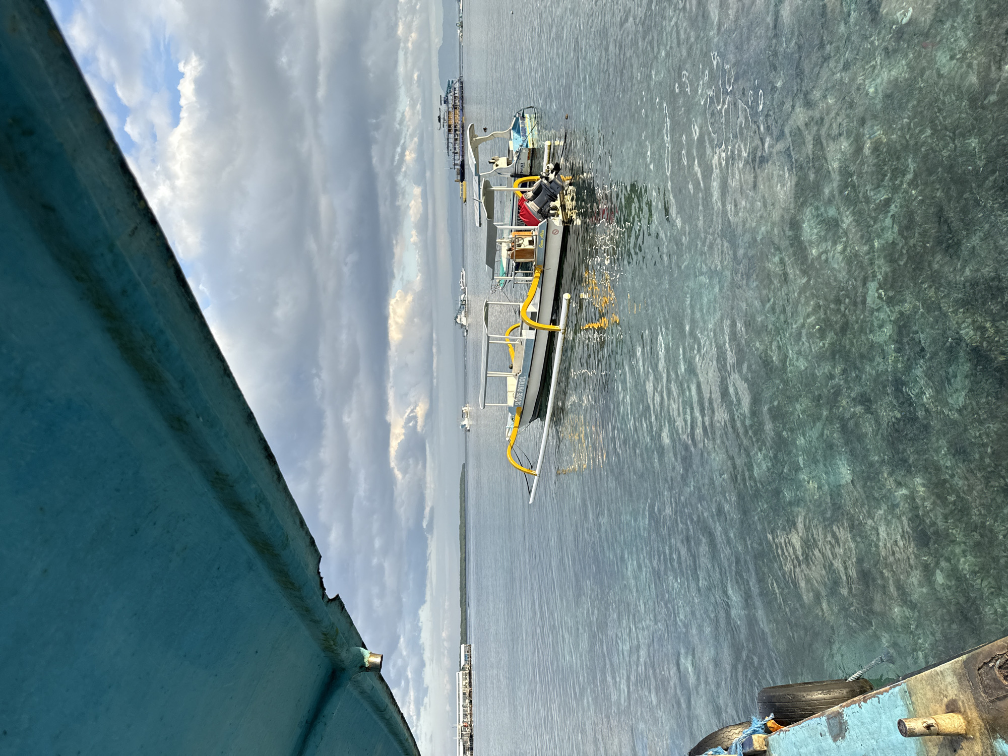 The image shows a boat in the water near Nusa Penida, Indonesia. The water is clear and turquoise. The sky is cloudy. There are other boats in the distance. The foreground shows part of another boat, with a blue painted exterior.