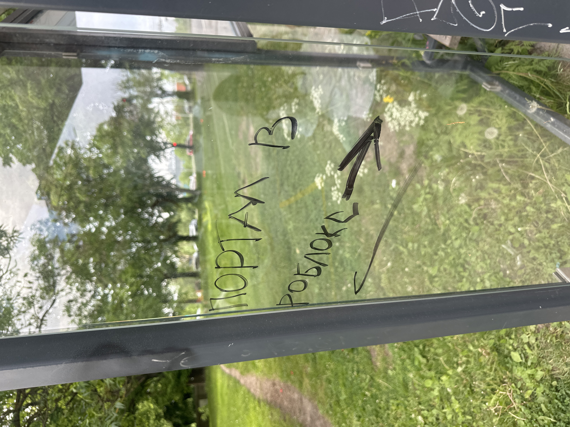 The image shows a glass pane, likely part of a bus stop shelter, with graffiti written in black marker. The text appears to be in Russian and reads "ПОРТАЛ В РОБЛОКС", which translates to "PORTAL TO ROBLOX". A crudely drawn arrow points to the right, indicating a direction. The background is blurred and shows greenery, trees, and a path, suggesting an outdoor urban environment. There are no people or visible interactions in the image. The graffiti is the primary subject.