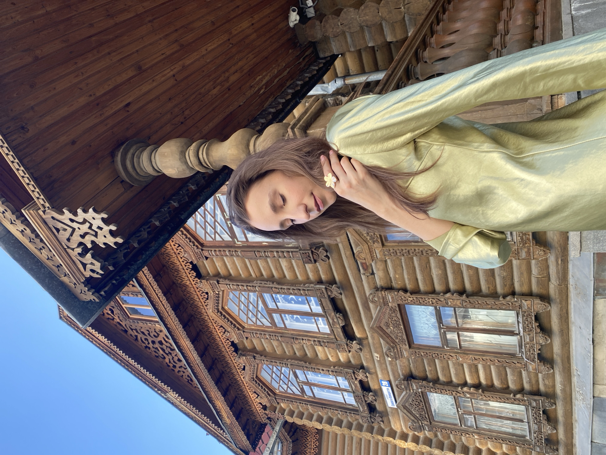A woman stands next to the Sevastyanov House in Yekaterinburg, Russia. She is wearing a shiny green jacket and has long brown hair. She has a flower-shaped ring on her finger and is touching her face. The Sevastyanov House is a wooden building with intricate carvings and ornate window frames. The sky is blue.