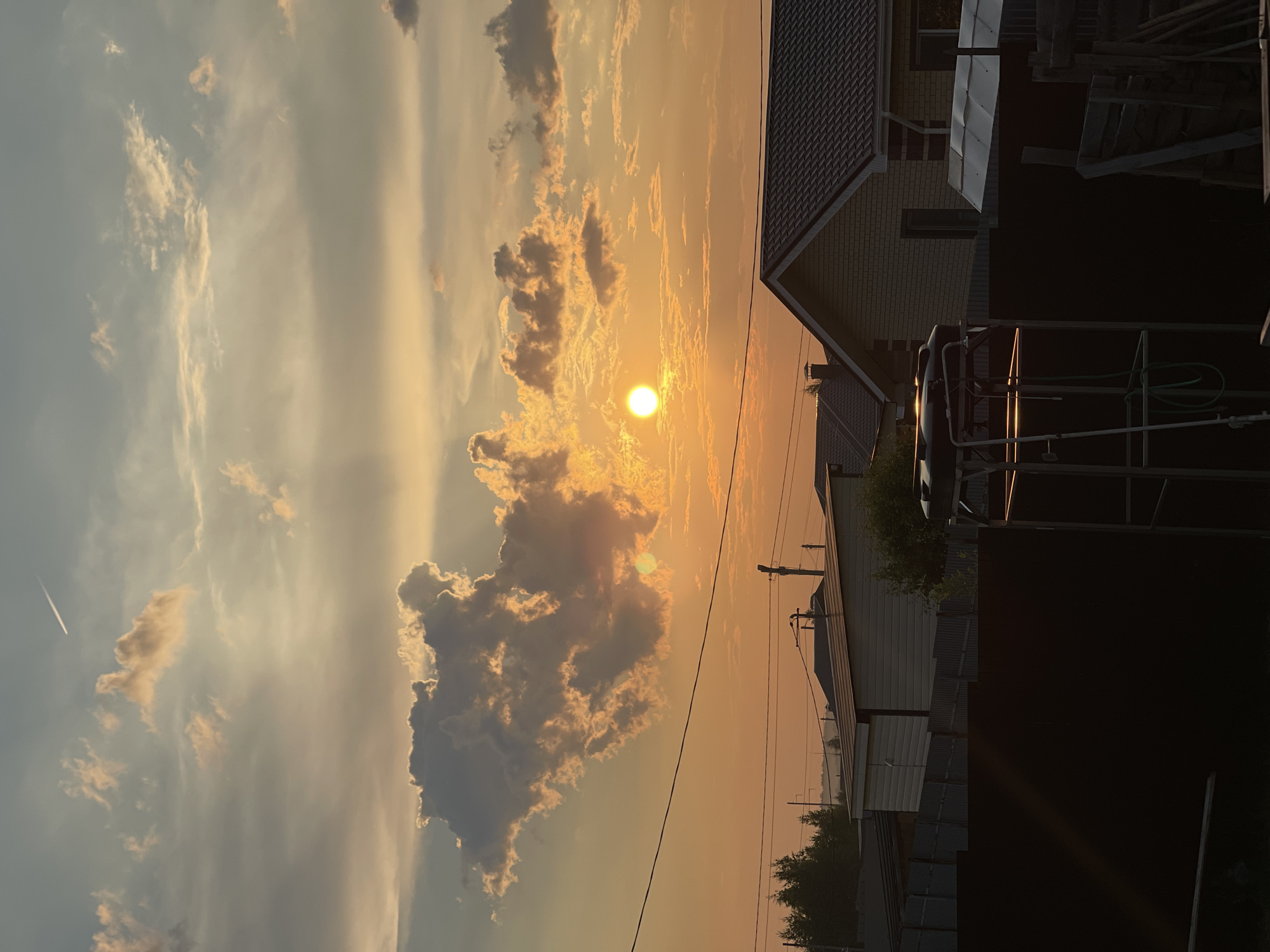 A sunset scene captures a sky filled with warm hues of orange and yellow, dotted with dramatic clouds. The sun, a bright orb, is partially obscured by a large, textured cloud formation in the upper center of the frame. A thin contrail from an airplane is visible in the far upper right corner. In the foreground, a dark fence stretches across the lower portion of the image. Behind the fence, residential structures with tiled roofs and brick exteriors are visible. A metal structure with some visible tubing stands in the right foreground, catching the light of the setting sun. Trees line the horizon in the middle ground. Power lines and utility poles are also present, traversing the sky. The overall impression is one of a tranquil evening in a suburban or rural setting.