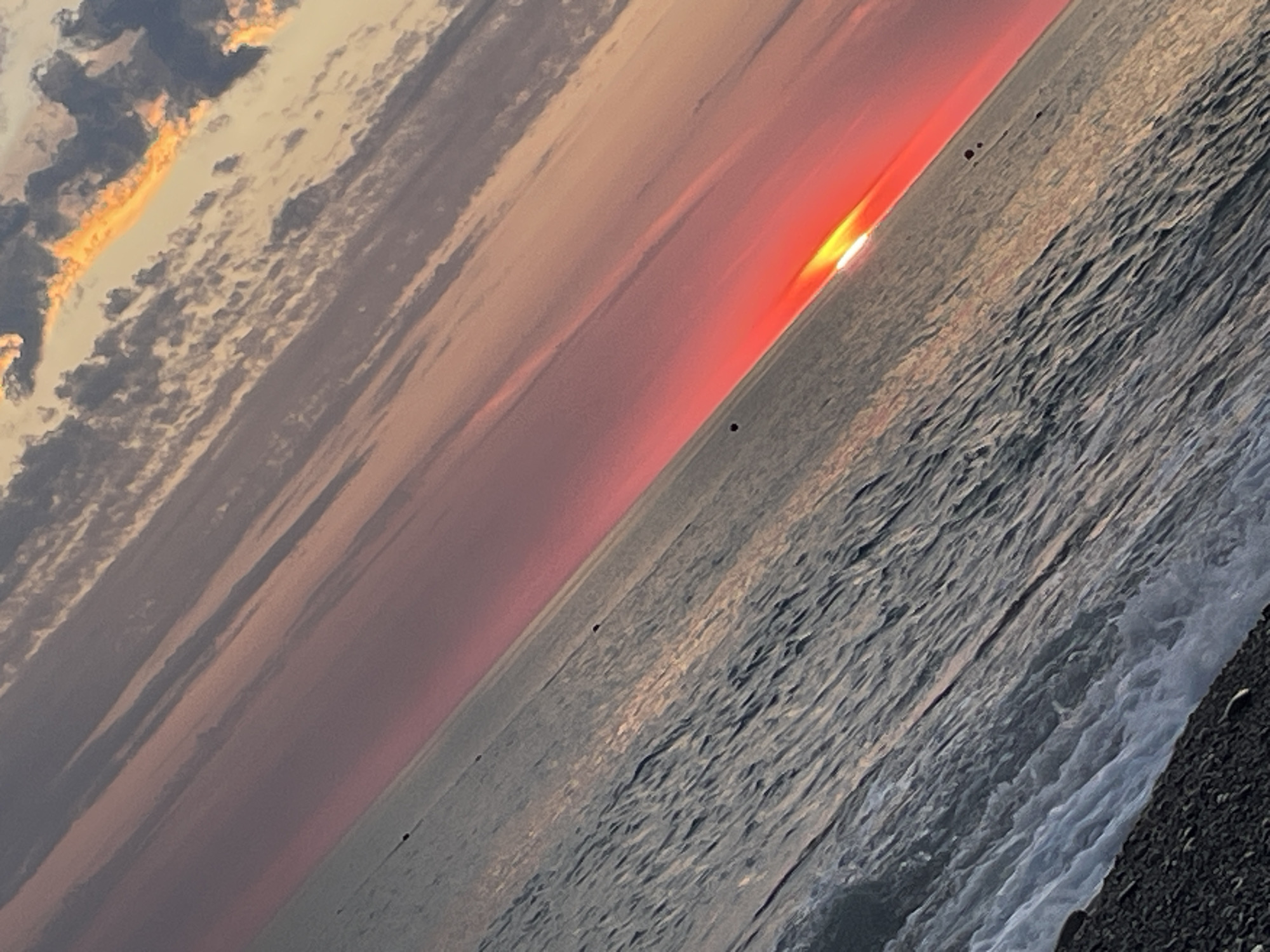 The image shows a sunset over a body of water, likely the ocean, with a pebble beach in the foreground. The sun is low on the horizon, casting a bright orange and red glow across the sky and reflecting on the water. The clouds are also tinged with orange and pink. The water is choppy, with small waves breaking on the shore. The image is taken at an angle, giving it a dynamic and dramatic feel. The location is unknown, as is the presence of any people or specific objects.