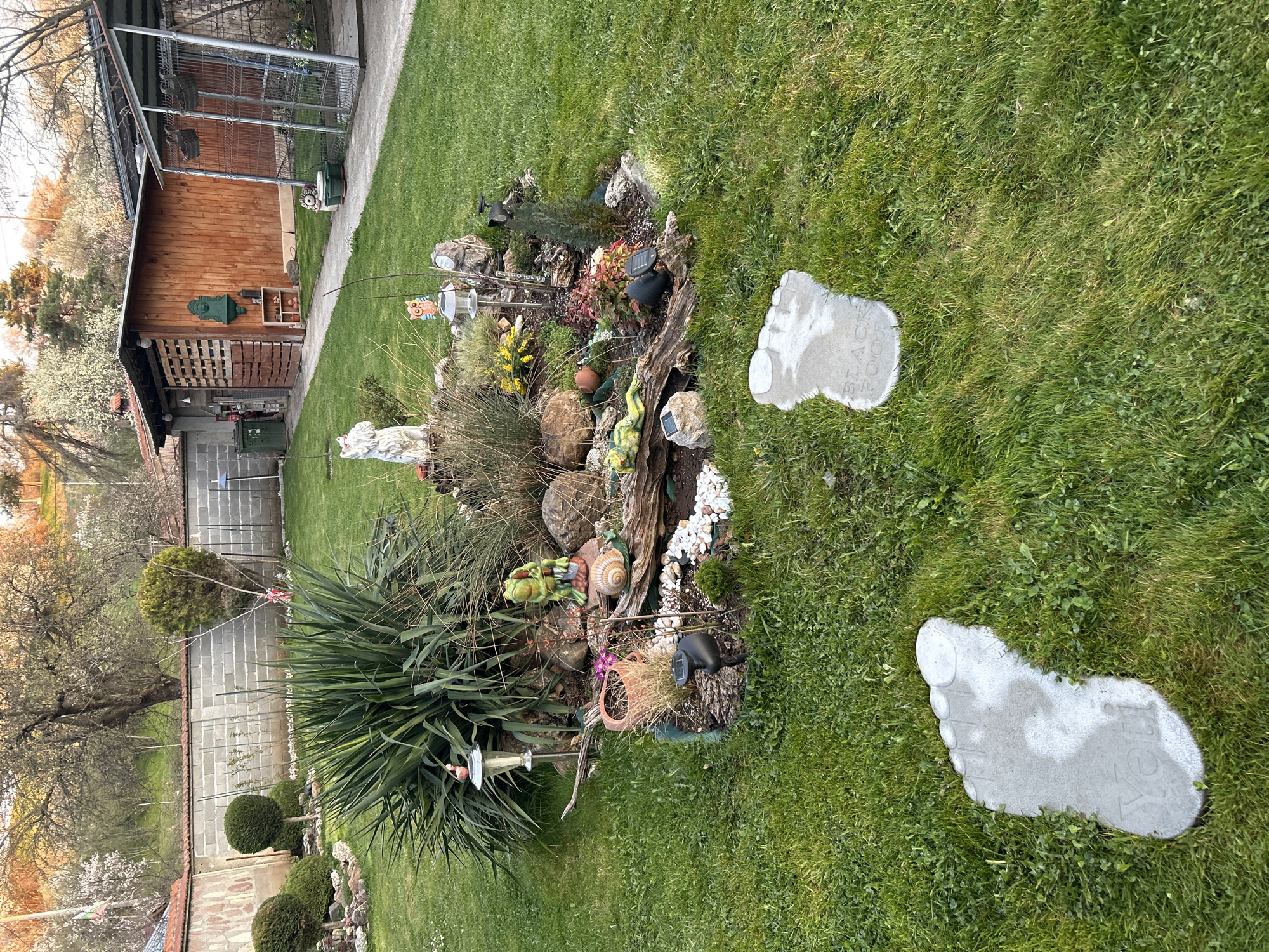The image shows a garden in Buhovo, Bulgaria. A wooden building with a metal enclosure stands to the left of the frame. The central focus is a landscaped rock and plant arrangement featuring various decorative elements like statues, snail shells, and solar lights. Two concrete stepping stones in the shape of bare feet are positioned on the lawn, leading away from the arrangement. The garden is well-maintained with neatly trimmed bushes along a wall in the background.