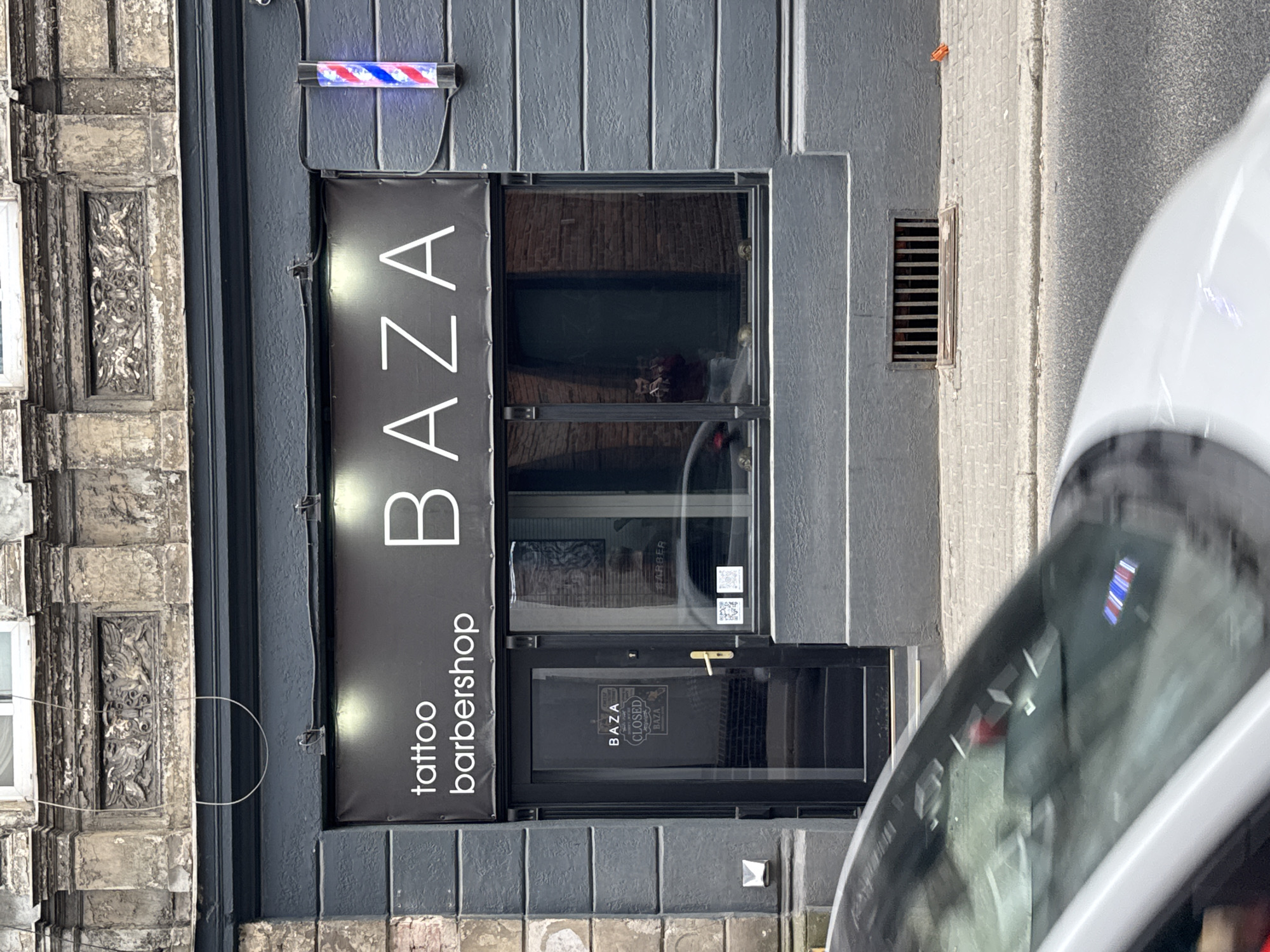 The image captures the front of "BAZA," a tattoo and barbershop located in Bydgoszcz, Poland. The shop's sign is prominently displayed above the entrance, featuring white lettering on a black background. A traditional barber's pole with red, white, and blue stripes is affixed to the wall above the sign. The entrance is a double-door style with a partially visible interior. The building's facade is characterized by textured, weathered stone and a gray painted wall. A metal grate is set into the sidewalk nearby. Part of a white vehicle, likely a car, is visible in the foreground on the right side of the frame.