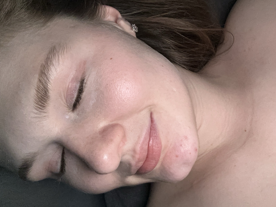 A close-up, angled portrait shot captures the left side of a fair-skinned individual's face, from the nose to the upper chest, located in the city of Kazan, Russia. The subject's eyes are closed, exhibiting long eyelashes and well-defined, light brown eyebrows. A subtle, relaxed smile is present on the subject's lips. The skin displays minor redness on the eyelids and several small, red blemishes on the chin. Brownish hair is visible near the subject's right ear, which features a reflective, possibly diamond-encrusted, stud earring. The bare neck and upper chest are partially visible. The background is indistinct and dark, indicative of an indoor, low-light environment.