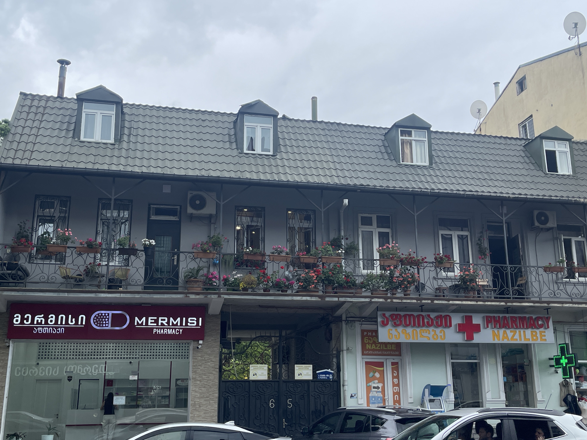 The image shows the facade of a multi-story building in Batumi, Georgia, on a cloudy day. The building features a gray, wavy-tiled roof with several dormer windows. The second floor has a balcony adorned with numerous potted flowers in various colors, including pink and red. Below the balcony, on the ground floor, are two pharmacies. The pharmacy on the left has a prominent red and white sign with "MERMISI PHARMACY" written in both Georgian and English. The pharmacy on the right has a red cross and "PHARMACY NAZILBE" written on its sign. The entrance to the building, located between the two pharmacies, is a large black metal gate with decorative scrollwork. A sign on the gate displays the numbers "65". Cars are parked in front of the building. The sky is overcast.