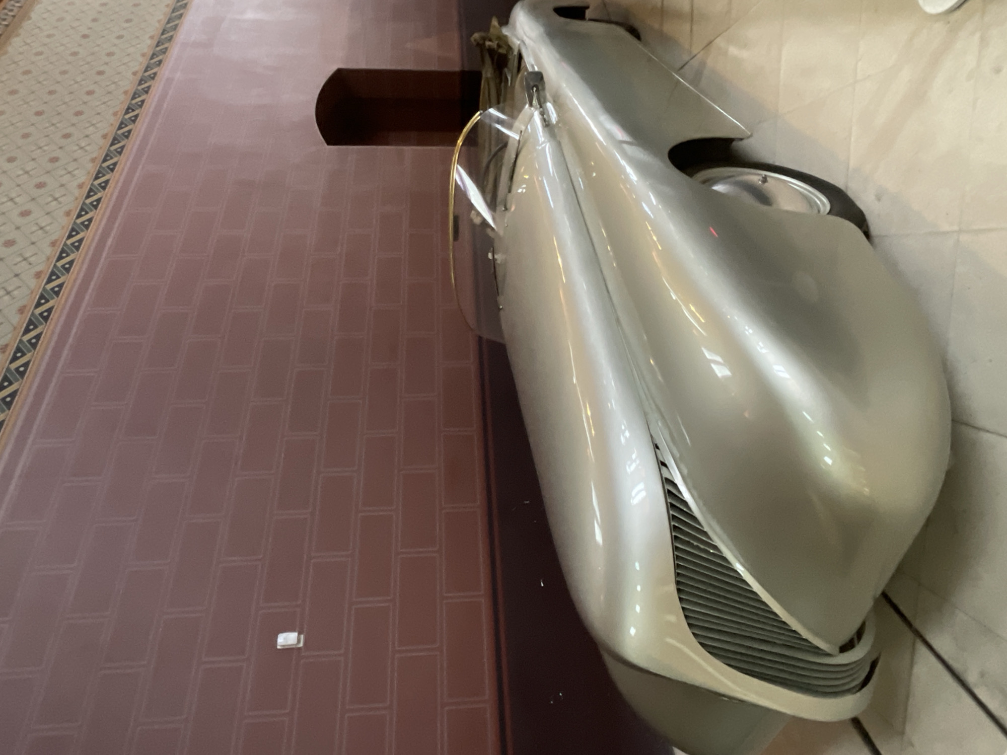 A sleek, silver convertible car with a streamlined design is parked indoors. The car has a gold trim around its windshield and what appears to be a brown fabric convertible top tucked behind the seats. The background features a brick-patterned wall and a decorative border near the ceiling, suggesting an interior space such as a museum or exhibition hall. The lighting highlights the car's polished surface. The car is positioned at an angle, showcasing its front grille and the curve of its hood. There are no people visible in the image. The car is positioned on a light-colored tiled floor. The architectural style of the background suggests a building with historical significance.