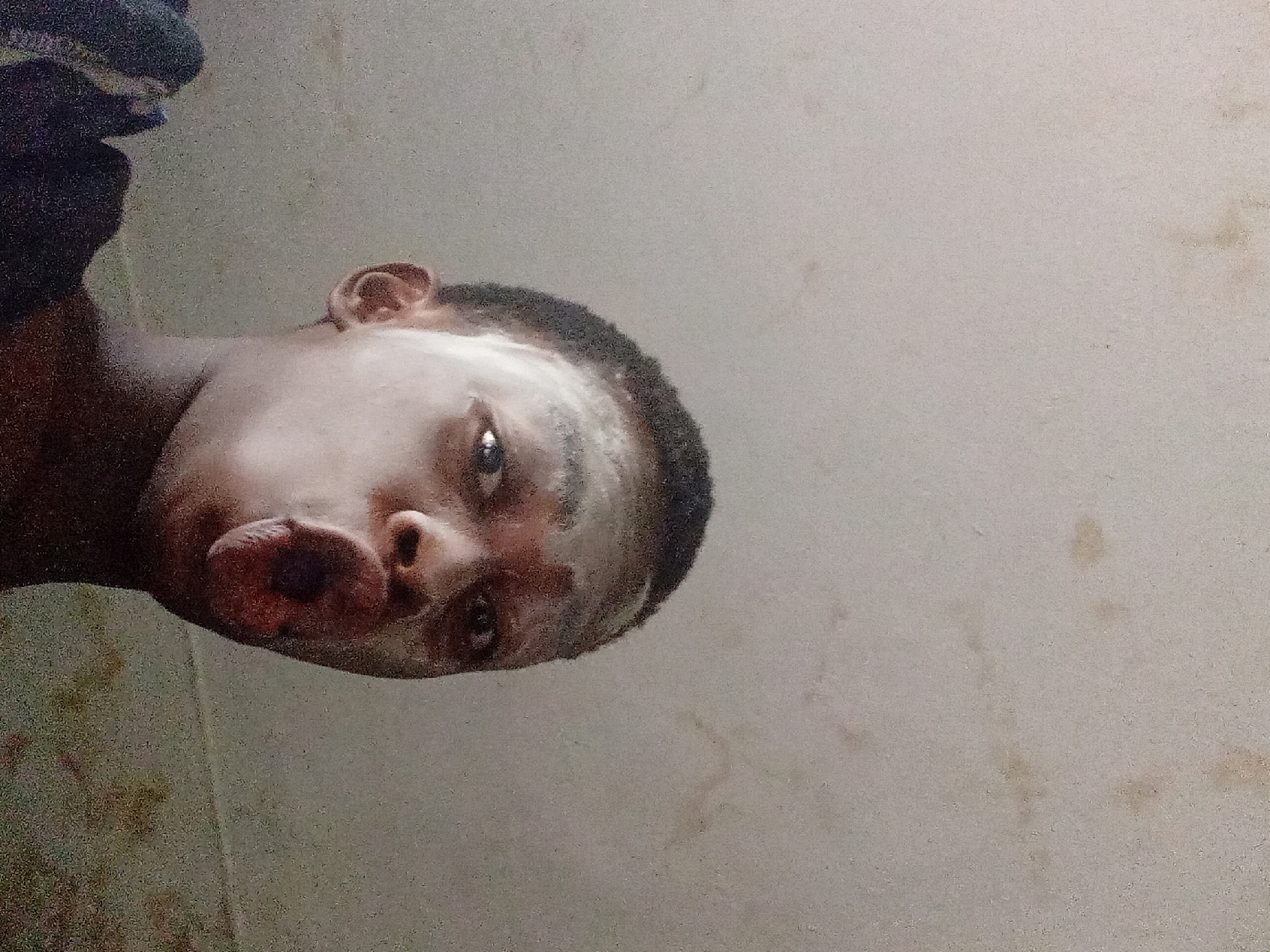 A medium shot photograph shows a young, dark-skinned person positioned at an unusual angle, with their head near the top and left side of the frame. Their face appears to be covered with a white substance, possibly a cream or mask. The background is a light, mottled wall with some stains. The image appears to have been taken indoors, but there are no other distinct features to indicate a specific location in Nkpor, Nigeria.