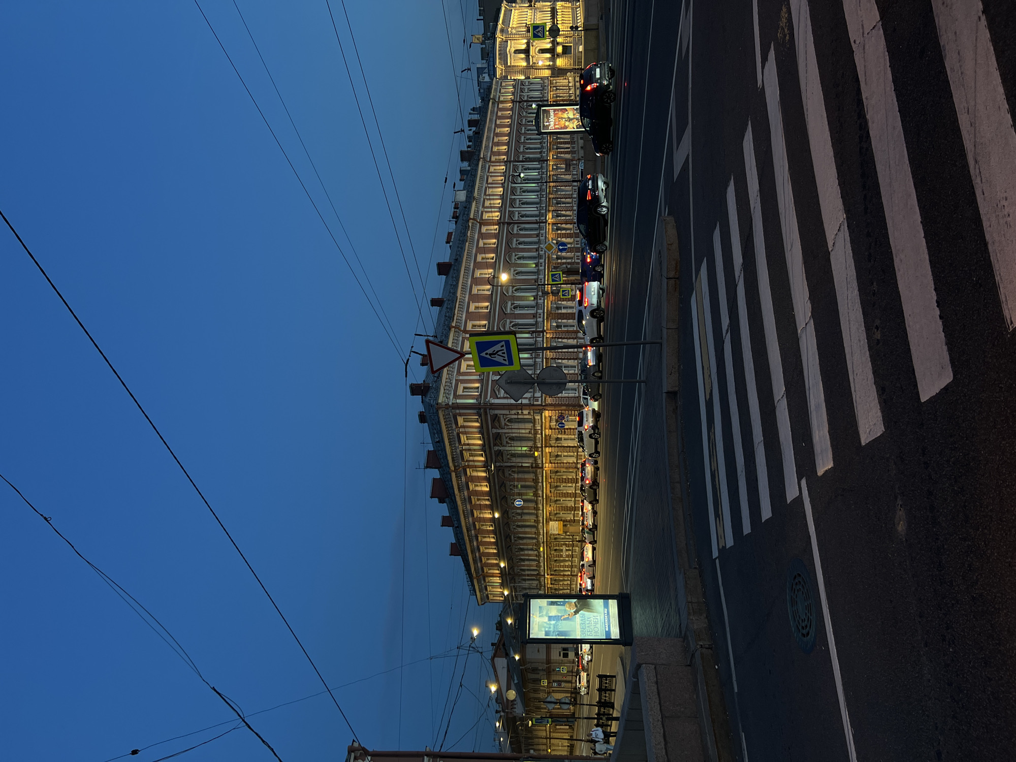 In the city of St. Petersburg, Russia, at dusk, a long, multi-story building with a reddish-brown facade is illuminated by warm lights. The building features rows of windows and decorative architectural elements. In the foreground, a pedestrian crossing is marked on the asphalt road. Several cars are visible on the street, some in motion and some parked. A digital billboard stands to the left of the building, displaying an advertisement. Overhead, power lines crisscross the dark blue twilight sky. The scene depicts a street view of an urban environment at the transition from day to night.