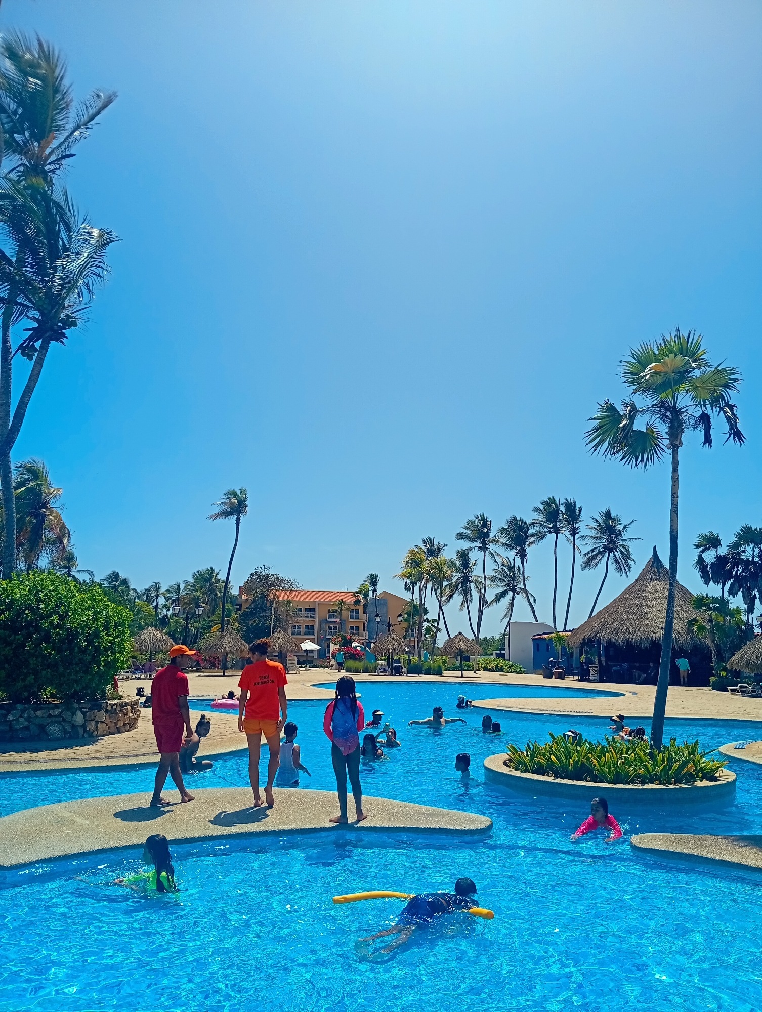 The image depicts a sunny scene at a resort in Playa El Agua, Venezuela. In the foreground, a large, interconnected swimming pool with clear blue water is filled with people swimming and playing. Several small, concrete islands are scattered throughout the pool. On one of these islands, two lifeguards wearing orange shirts and shorts are walking alongside a young girl with a pink backpack. Various palm trees and thatched-roof structures surround the pool area. A tan-colored building is visible in the background.