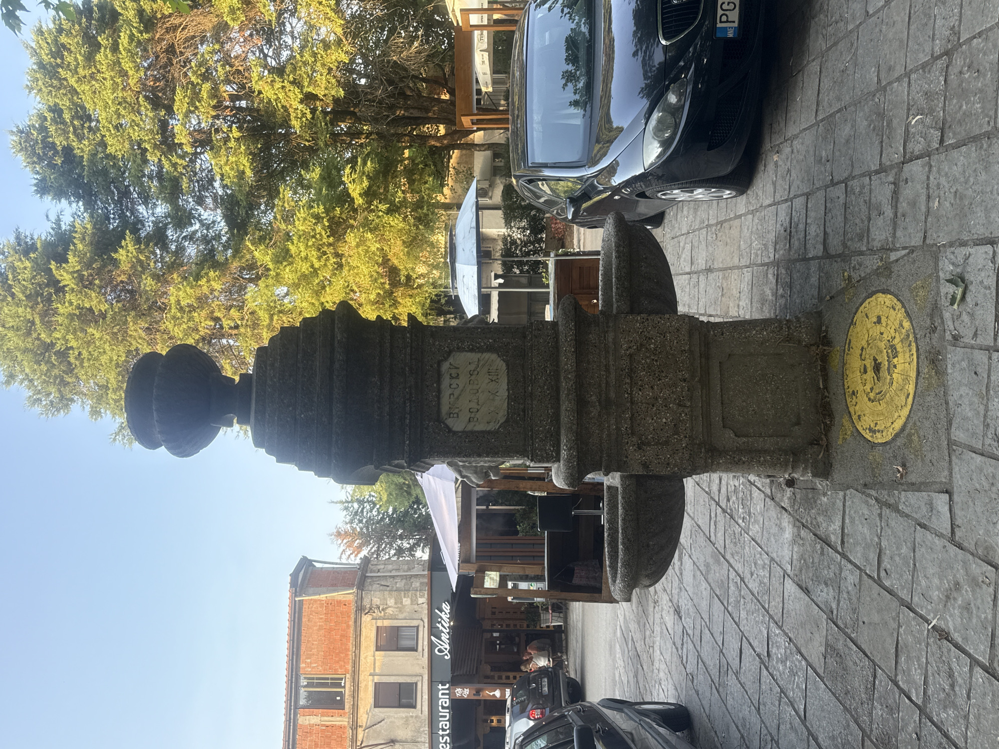 In the city of Virpazar, Montenegro, a stone fountain stands prominently in a paved courtyard. The fountain features a carved inscription that reads "ВИР СКИ" on the top line and "ВОДОПР" on the second line, followed by "XX XIII" on the third line. Two basins protrude from the sides of the fountain. To the right of the fountain, a black car is parked, and a yellow circular manhole cover is visible on the ground in front of the fountain. In the background, several buildings and trees line the street, with awnings and umbrellas suggesting outdoor dining areas. A few people are seated at tables outside a restaurant labeled "Antika." The sky is clear and blue.