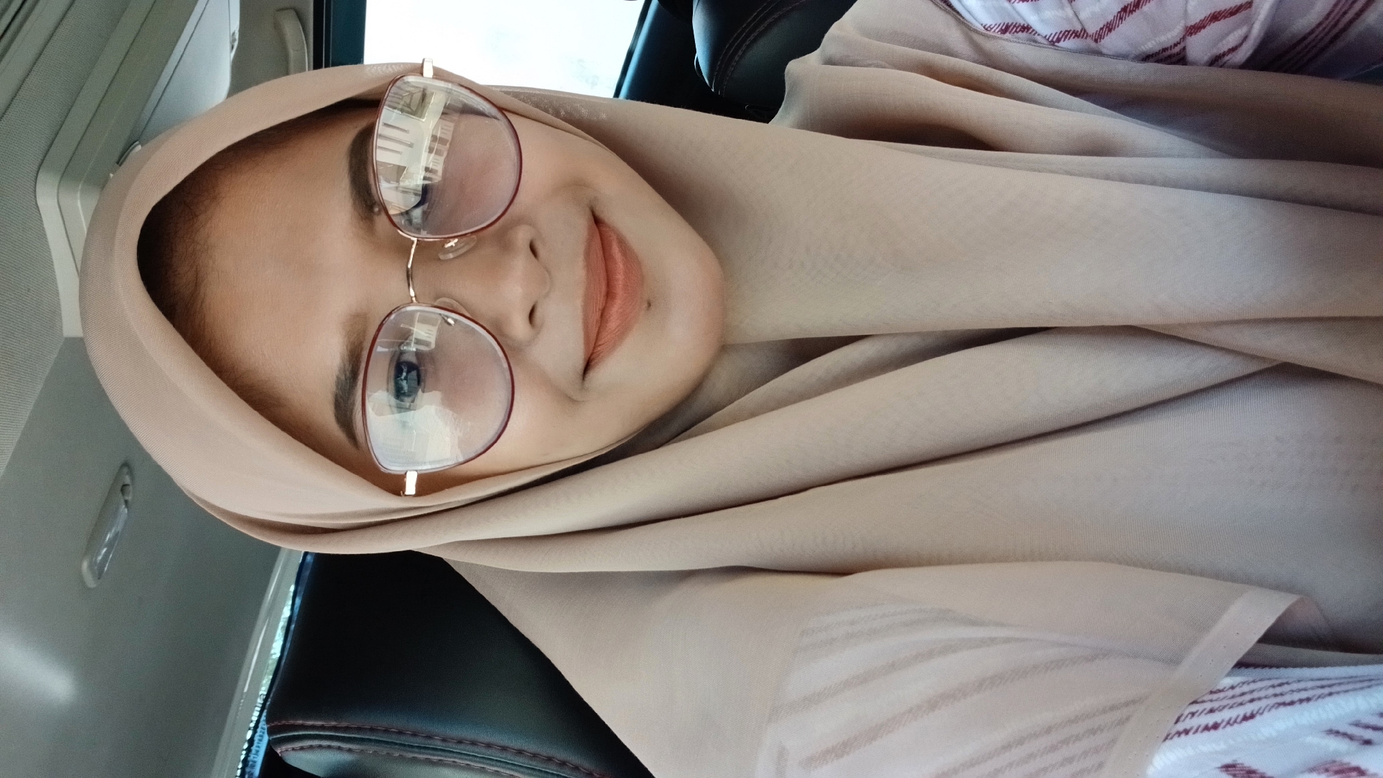 A woman is posing for a selfie in what appears to be the interior of a car. She is wearing a light beige headscarf (hijab) and round glasses with a gold-colored frame. She has fair skin and is smiling slightly. The seat beside her is dark with red stitching. The location is difficult to determine with certainty, however, based on the prompt, it is presumed to be within Lhokseumawe, Indonesia.