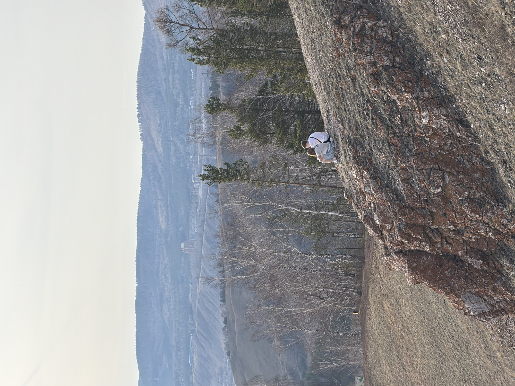 The image depicts a rock climber clinging to a rock face. The person is wearing a white shirt, and a harness, and appears to be focused on their ascent. The rock face is textured and rugged, suggesting a natural outdoor setting. In the background, a hazy landscape with trees and distant buildings is visible. The location is the Stolby Nature Sanctuary near the city of Krasnoyarsk, Russia.