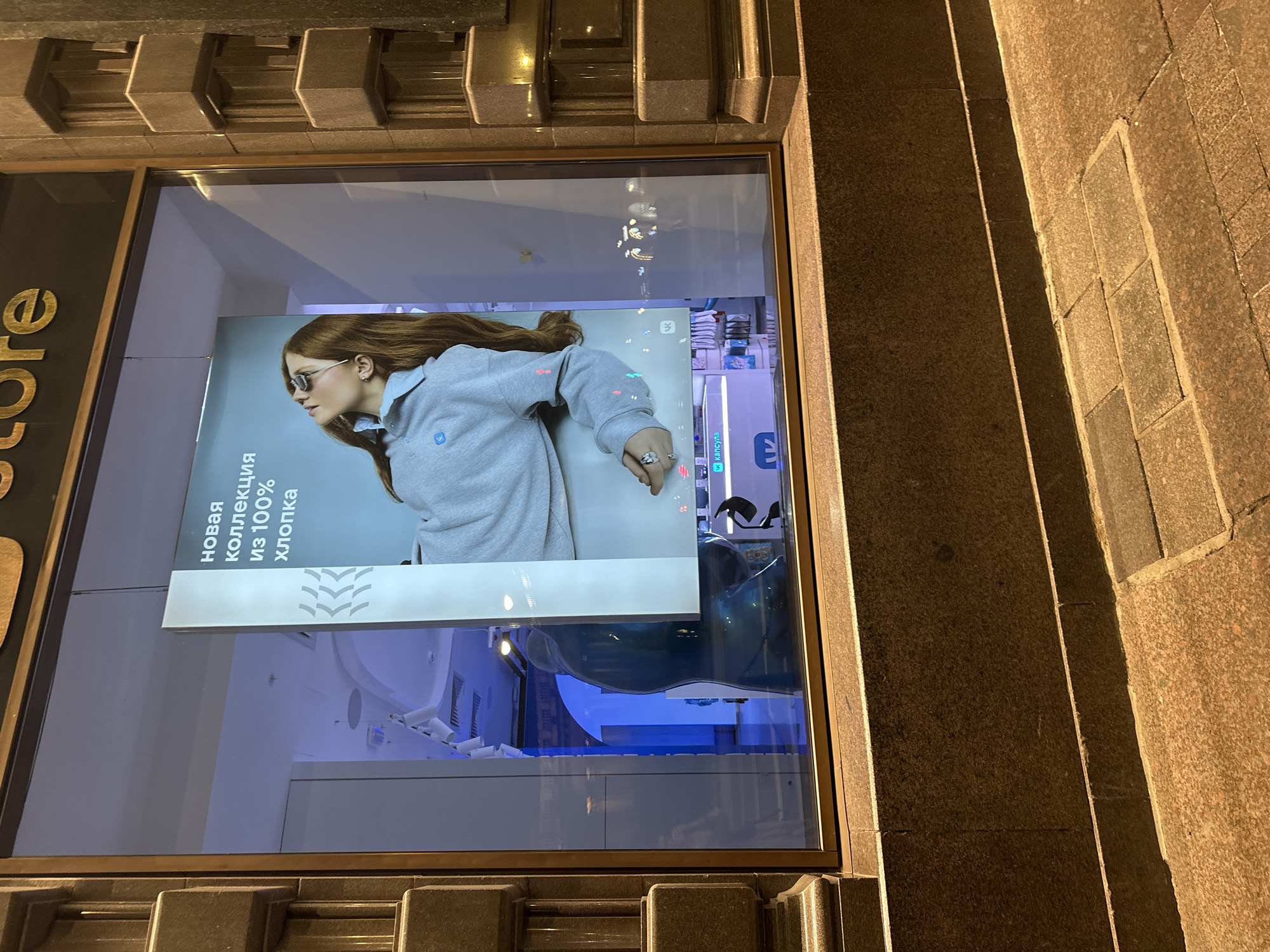 The image shows a window display in Saint Petersburg, Russia. Inside the window is an advertisement featuring a woman wearing a gray hoodie and sunglasses. The text on the advertisement is in Russian and mentions a "new collection" made of "100% cotton." The display appears to be in a store with bright blue lighting. The exterior of the building is made of stone, and the sidewalk is paved with brown tiles.