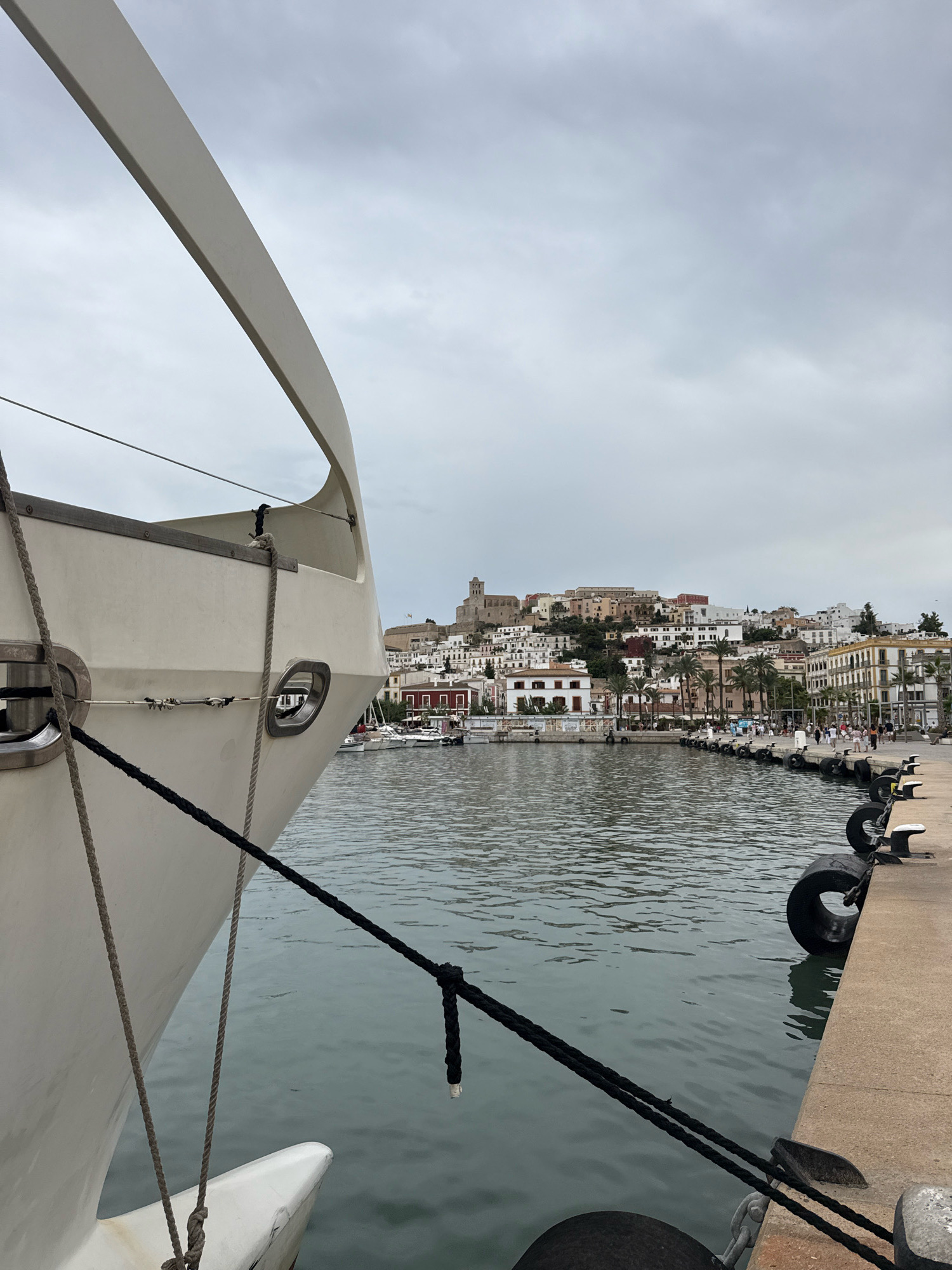This is a photograph taken from a docked boat, looking towards the city of Ibiza, Spain. The foreground is dominated by the side of a white boat, with ropes securing it to the dock. In the middle ground, the water of the harbor is visible, with several other boats docked along the harbor wall. The background features a densely built cityscape climbing up a hill, characteristic of Ibiza's old town, Dalt Vila. The buildings are a mix of white and pastel colors, with a prominent church structure visible at the summit of the hill. The sky is overcast, casting a soft, diffused light on the scene. Several people are visible walking along the harbor promenade.