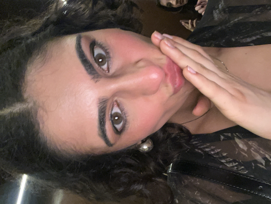 A close-up image captures a young adult female with dark, curly hair and dark brown eyes, directly engaging the viewer. She is performing a "blowing a kiss" gesture, indicated by her pursed lips and her right hand brought up to her mouth, palm slightly cupped. She wears large, silver-toned, dome-shaped earrings and a sheer, dark-colored patterned top. A thin necklace is visible around her neck. Her fingernails are manicured with a light polish. Her face shows applied makeup, including eyeliner and blush. In the blurred background, towards the lower right, the upper portion of a second individual's face and their raised right hand are visible. The setting is dimly lit and indistinct, suggesting an indoor environment with artificial lighting. The specific location within Paris, France, cannot be determined from the visual information present.