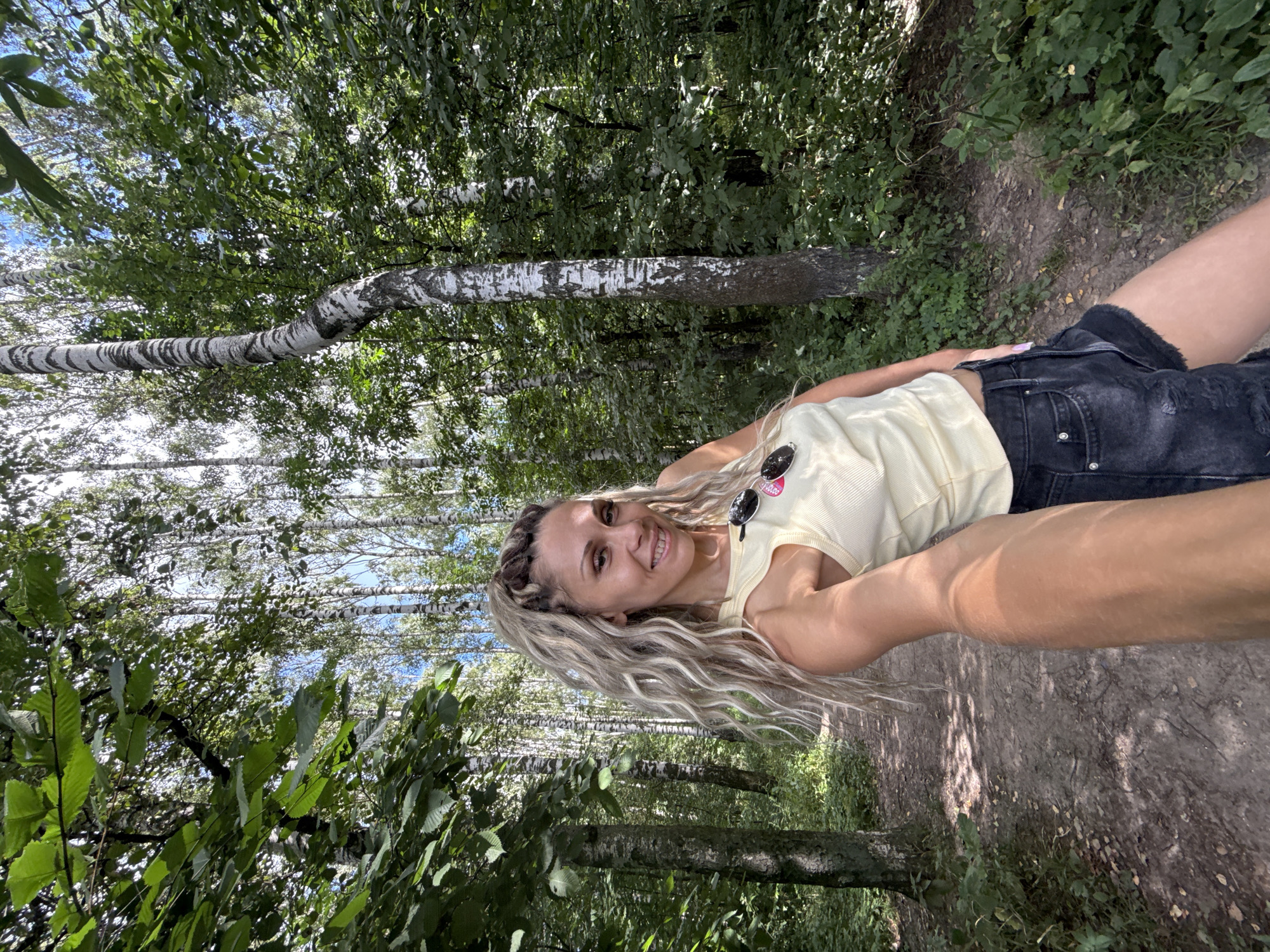 A woman is taking a selfie in a forest with birch trees. She is wearing a light yellow tank top and dark denim shorts. She has long, wavy blonde hair and is smiling at the camera. Sunglasses are clipped to her tank top. The setting is a dirt path through a wooded area with dappled sunlight filtering through the leaves. The trees appear to be primarily birch with their distinctive white bark. The sky is visible through the canopy, suggesting a bright, clear day. The overall mood is casual and relaxed, indicating an outdoor activity or a moment of leisure. The photograph was taken in Rusanovo, Russia.
