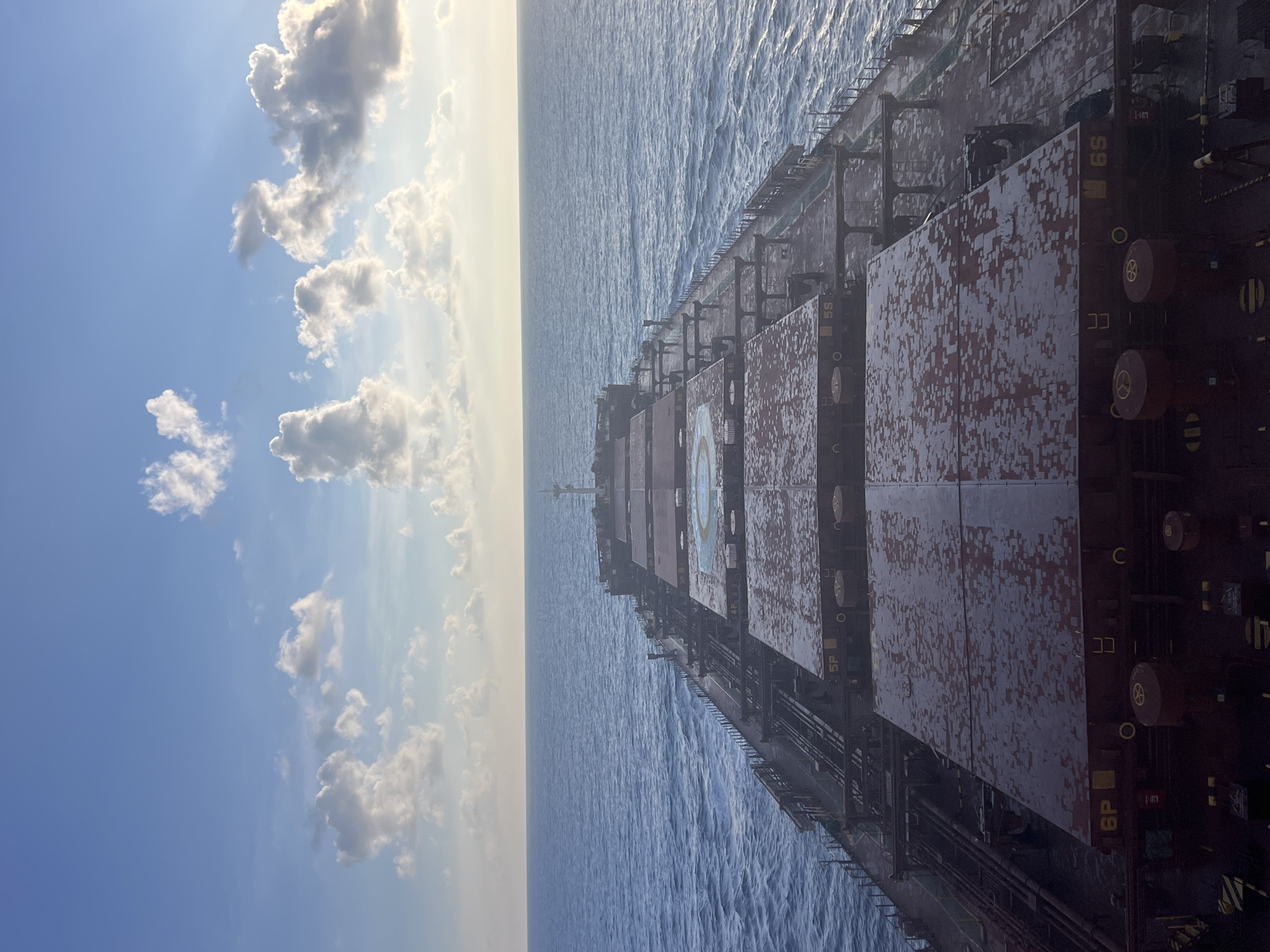 This is an aerial, high-angle, eye-level shot of the deck of a large cargo ship sailing on a calm, blue sea under a bright, cloudy sky. The ship's deck is the primary subject, with large, weathered, red cargo hatches filling the foreground and mid-ground. Many of these hatches have a speckled pattern of white and gray, possibly indicating frost or accumulated residue. The hatches are numbered, with "6P" and "5S" visible. Various pipes, railings, and equipment are visible along the sides of the deck. In the distance, the ship's superstructure, including a mast, is visible, leading to the horizon where the ocean meets the sky. The sky is a vibrant blue, with scattered white, puffy clouds, some of which are illuminated by the sun, creating a sense of depth and a soft glow. The sea is relatively flat with small ripples, reflecting the light from the sky. The overall scene conveys a sense of open space and maritime activity.