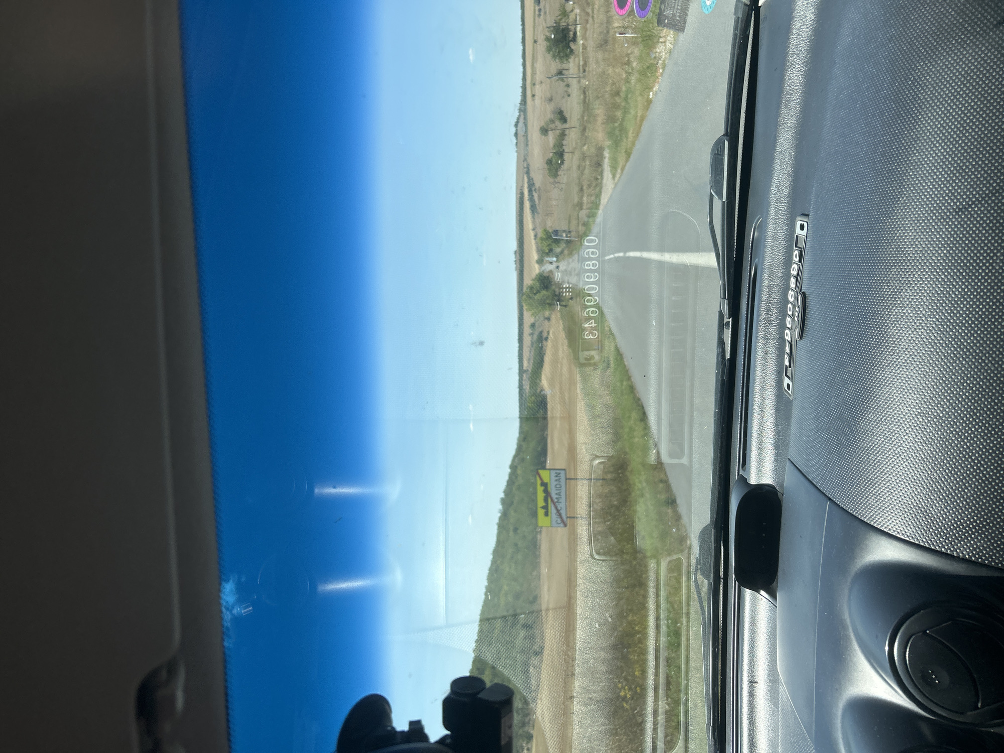 The image shows a view from inside a vehicle looking out through the windshield at a rural road in Cioc-Maidan, Moldova. The road stretches ahead into a landscape of dry fields and scattered trees under a clear blue sky. A yellow road sign with an image of a town and a red circle with a diagonal line through it, indicating the end of a settlement, is visible on the right side of the road, with the text "CIOC MAIDAN" below it. In the foreground, the dashboard of the vehicle is visible, including a dashboard camera mounted on the windshield and parts of the car's interior. The overall scene depicts a journey through the Moldovan countryside on a sunny day.
