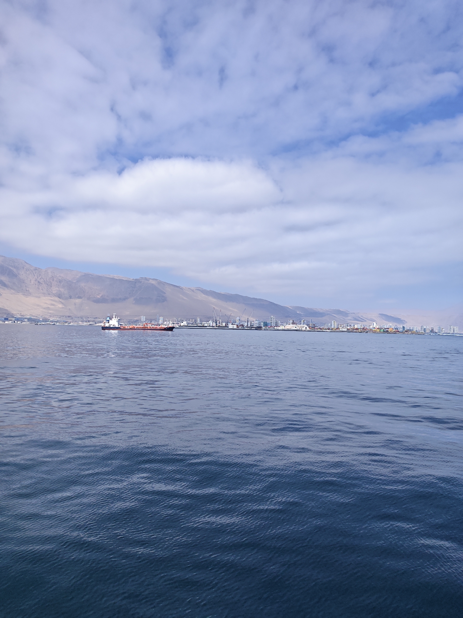 This is a wide shot of the bay of Iquique, Chile, as seen from the water. In the foreground, the dark blue water has small ripples across its surface. In the middle ground, a large cargo ship with a red hull and white superstructure is sailing to the left. In the background, a long line of buildings stretches across the coast, backed by barren, brown hills under a cloudy sky. The buildings appear to be a mix of residential and industrial structures, with cranes visible near some of the ports. The overall impression is one of a busy port city nestled against a desert landscape.