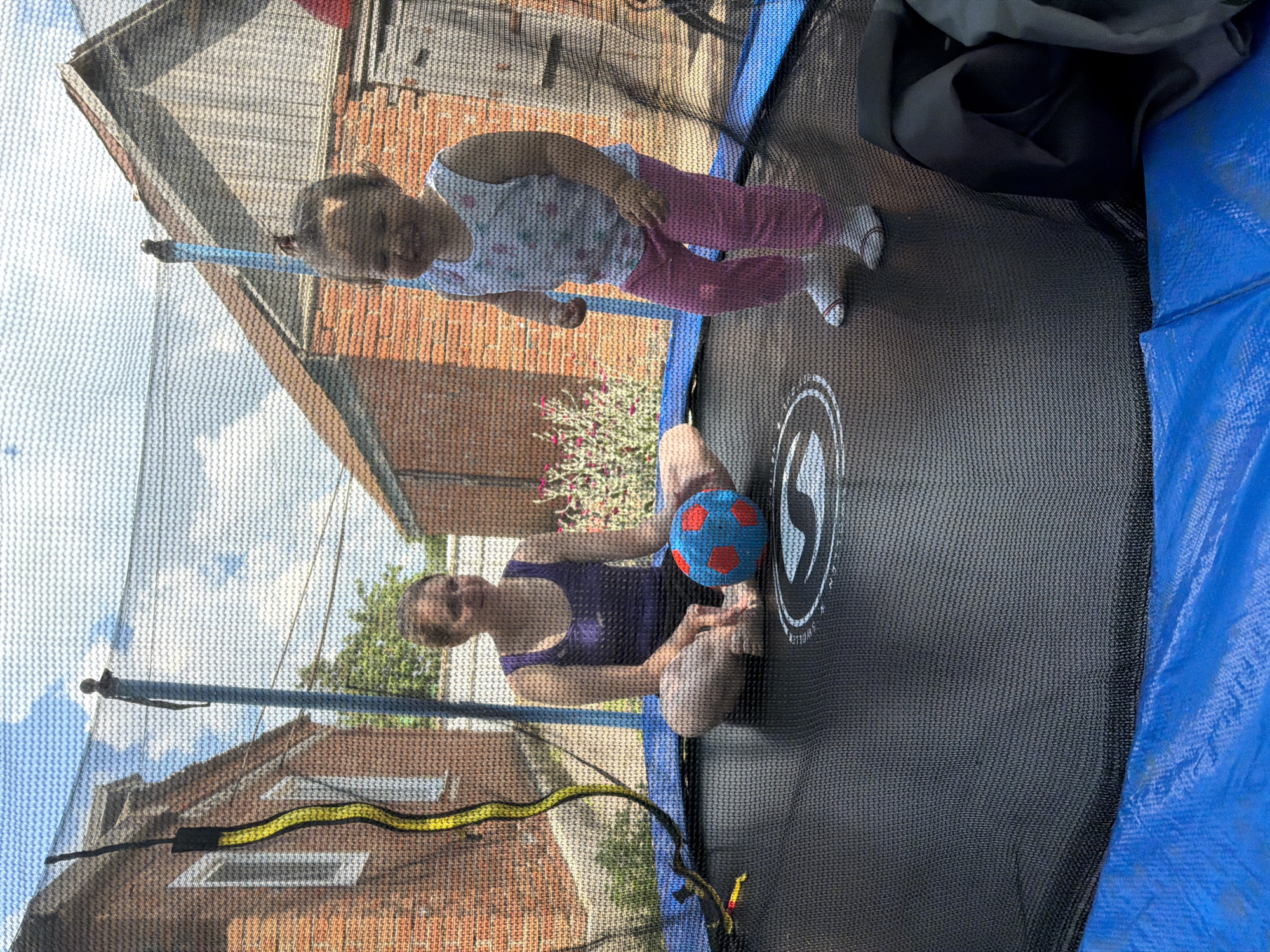In Komsomolskiy, Russia, a woman and a toddler are on a trampoline. The woman, seated cross-legged, wears a dark purple tank top and dark shorts. A blue and red ball rests between her legs. The toddler, standing, is dressed in a white floral tank top and pink pants, with white socks. They are in a backyard, with brick houses visible in the background. The sky is blue with scattered clouds.