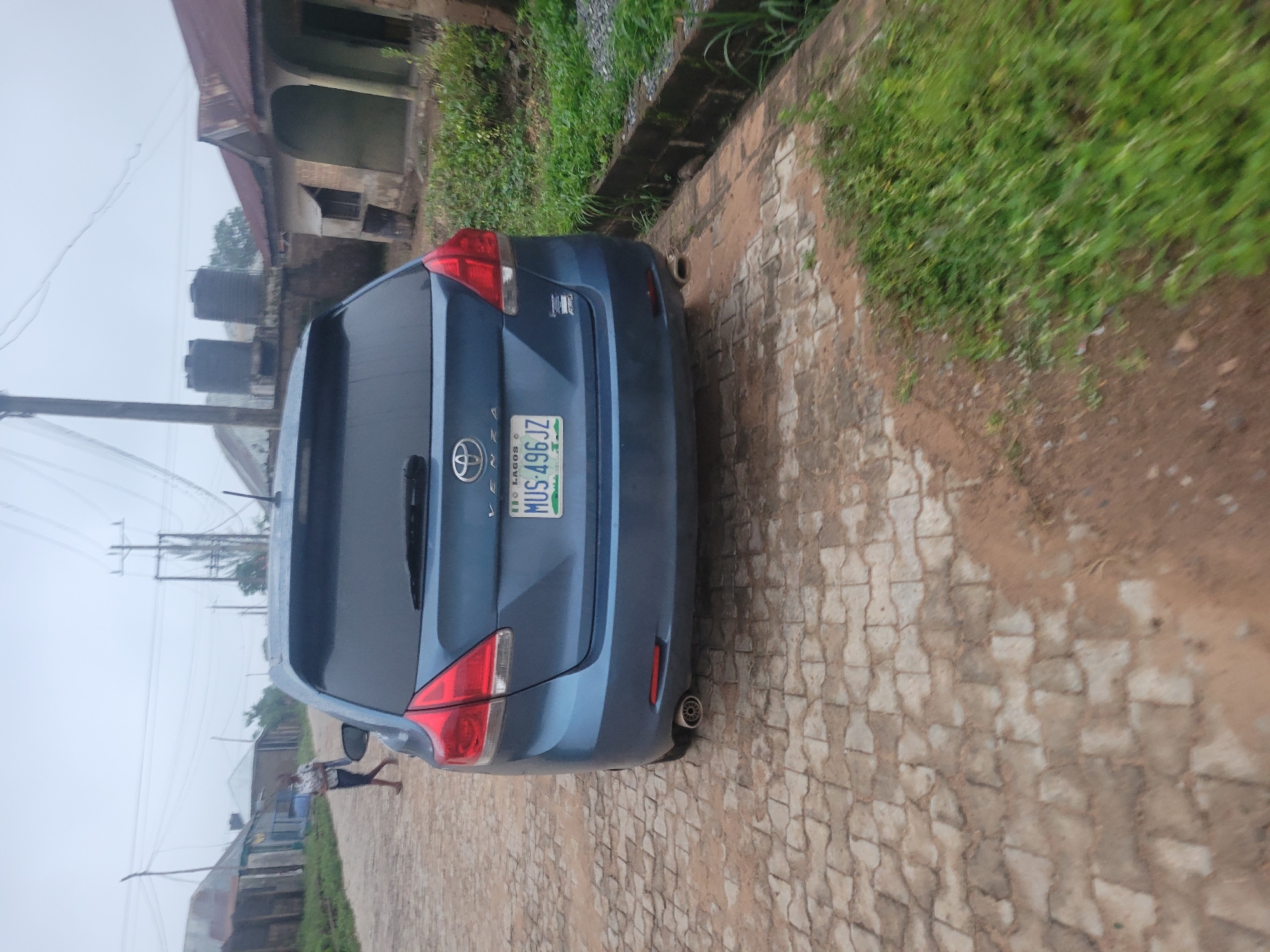 The image captures the rear view of a blue Toyota Venza parked on a cobblestone path in a seemingly rural or suburban area. The license plate reads "MUS-496JZ" and has "LAGOS" printed above the number. To the left of the car, a person is walking away from the camera. In the background, there are buildings with corrugated metal roofs, some water tanks, and utility poles with numerous wires. Overgrown green vegetation lines the right side of the path. The sky is overcast, indicating a cloudy day.