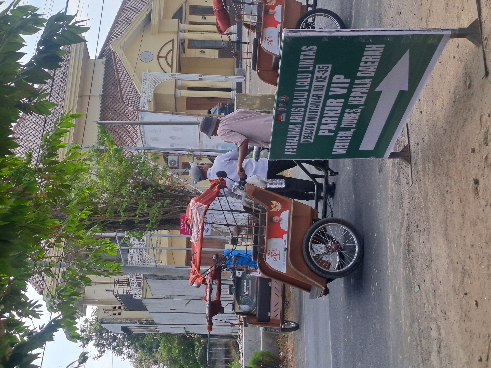 The image depicts a street scene in Mojokerto, Indonesia. In the foreground, a green sign with white text and an arrow points to the right, indicating "PARKIR VIP MENTERI, DUBES, KEPALA DAERAH," which translates to "VIP PARKING MINISTERS, AMBASSADORS, REGIONAL HEADS." Two men are present near a brown pedicab, one wearing a light-colored cap and shirt, and the other a hat and a light-colored long-sleeved shirt. The pedicab has orange coverings and appears to be stopped on the side of the road. In the background, a building with a tiled roof and yellow facade is visible, along with trees and a fence. The overall environment suggests an outdoor, public space.