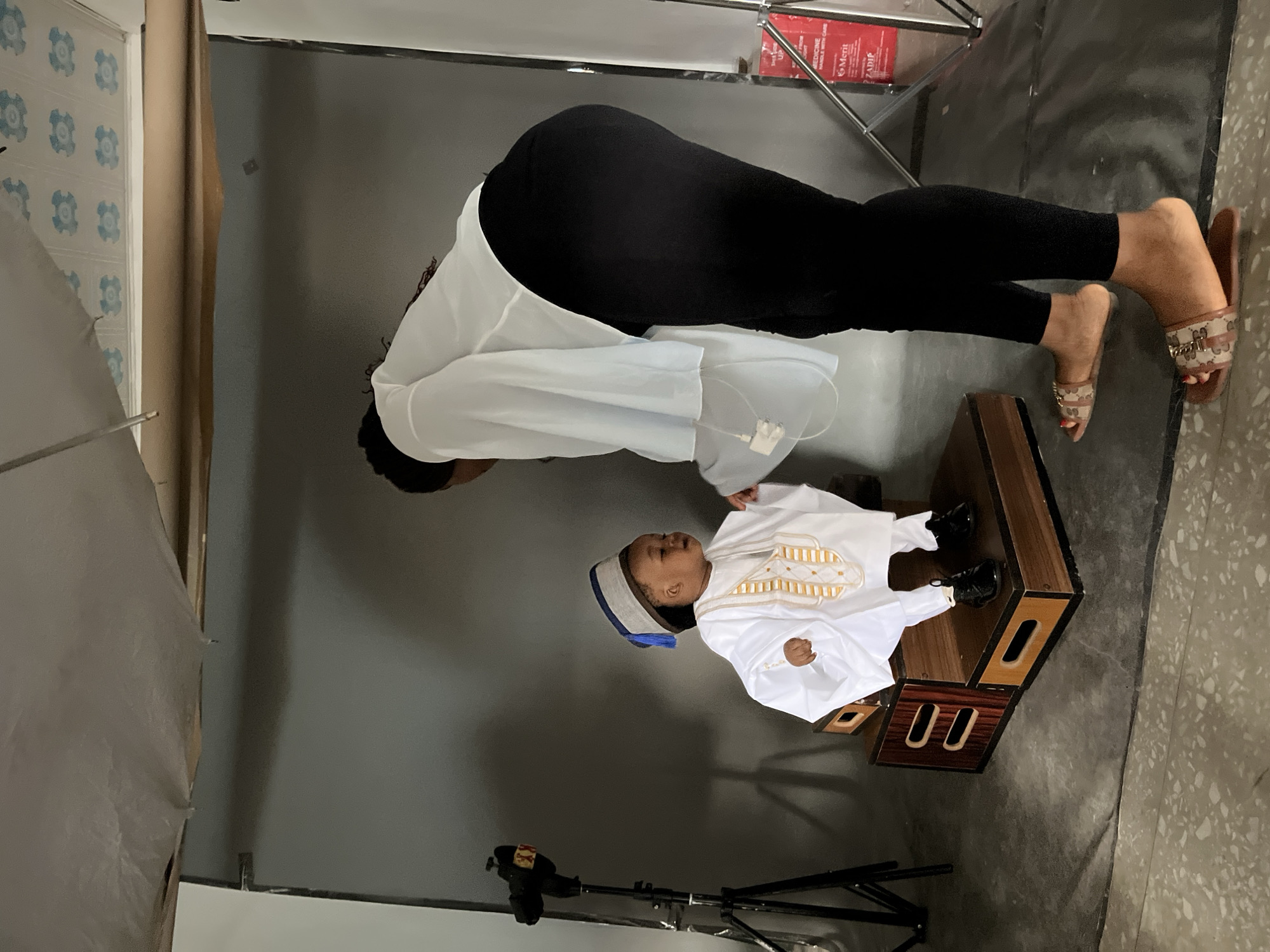 The image depicts a studio setting with a child dressed in traditional Nigerian attire. The child, a young boy, is seated on a wooden platform, wearing a white kaftan with gold embroidery and a blue and grey cap. A woman, presumably a photographer or assistant, is standing to the right of the child, leaning down and appearing to adjust the child's clothing. The background is a grey studio backdrop, with a lighting stand visible on the left. The floor is a textured concrete. The presence of a studio backdrop and lighting equipment suggests a professional photoshoot is taking place. There is no specific visual information within the image that indicates the location is Gura Top, Nigeria.