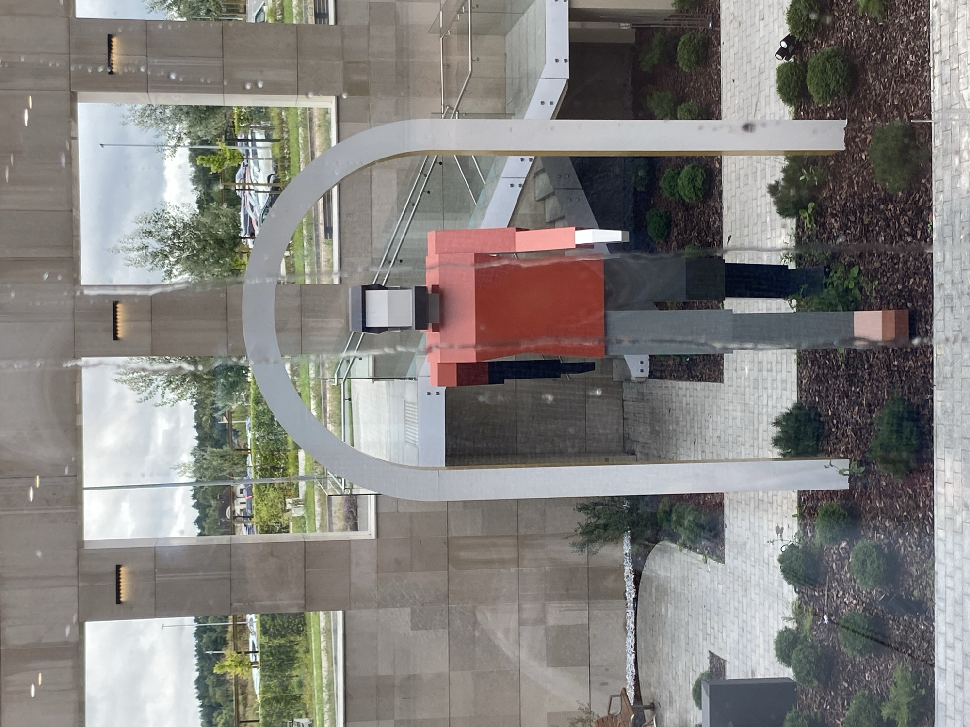 The image shows a large, abstract sculpture of a person made of rectangular blocks, predominantly red and grey, standing in an outdoor courtyard. The sculpture is framed by a large, white archway. Behind the sculpture, a modern building with large windows and stone facade is visible, as well as an outdoor staircase with glass railings. Beyond the building, a parking lot with several cars and a line of trees and greenery can be seen under a cloudy sky. The foreground features a paved courtyard area with small, manicured bushes and dark mulch. The image appears to be taken through a window, as evidenced by reflections and water droplets on the glass.