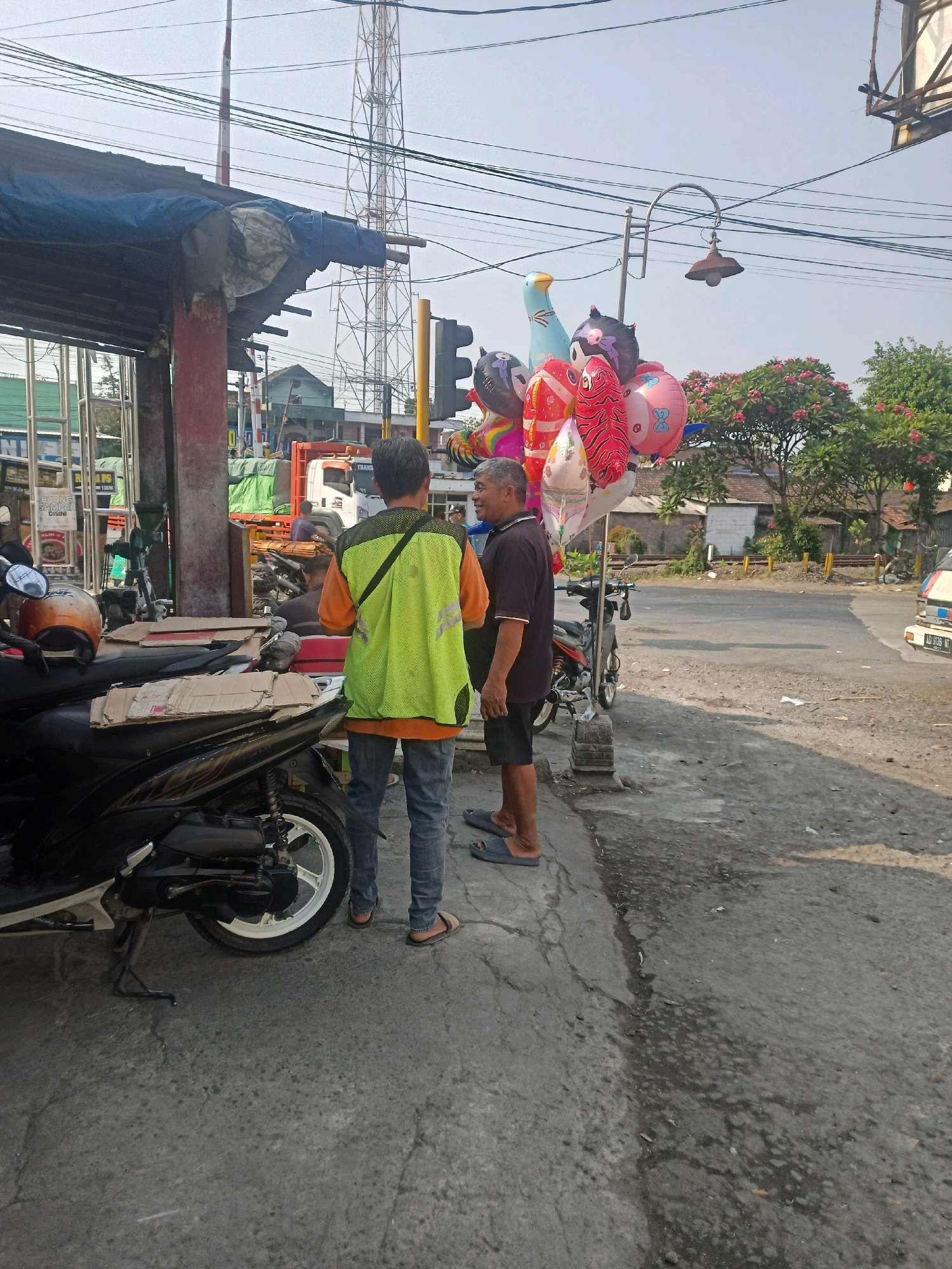 The image depicts a street scene in Ngandul, Indonesia, likely near a roadside vendor. Two men are standing on a cracked asphalt surface next to a motorcycle. The man on the left is wearing an orange shirt and a bright green reflective vest. He is looking towards the right, towards the other man. The man on the right is wearing a dark polo shirt and dark shorts. He is also looking to his right, and appears to be smiling.
In front of the men, a collection of colorful balloons are displayed, some shaped like animals and characters. The balloons are attached to a stand which is propped up by a concrete block. Behind the men, there is a building with a blue tarp covering its roof and a red support column. A traffic light is visible on a pole to the left of the balloons. A truck with a green tarp on its load is parked in the background. Further in the distance, trees with pink blossoms are visible, as well as power lines stretching across the sky. A white car with an Indonesian license plate is parked on the right side of the street. The overall impression is of a typical daytime scene in a bustling Indonesian town.