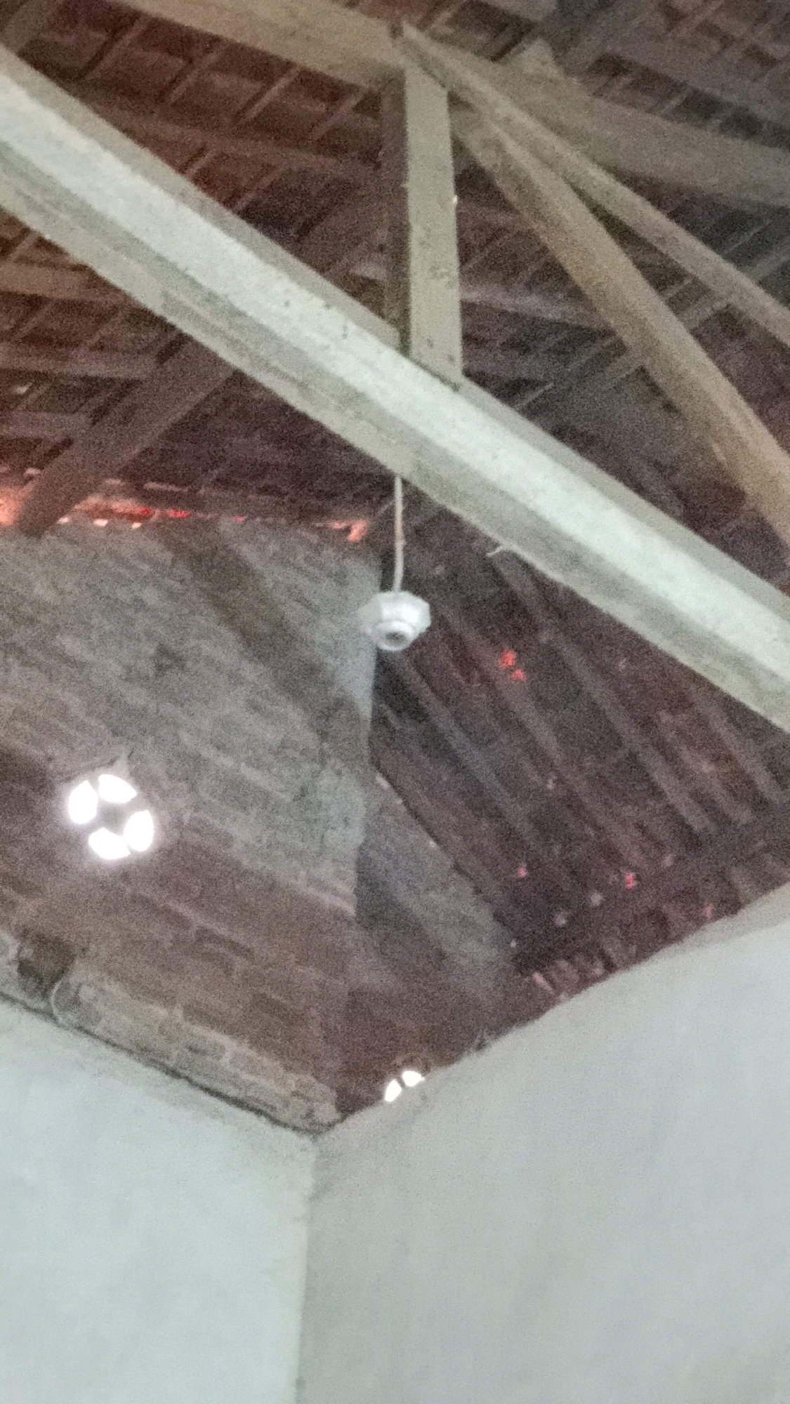 The image shows the interior of a building, likely a barn or an old structure, with a wooden roof and exposed beams. A small, white security camera is hanging from a wire in the center of the frame. The walls appear to be made of stone or brick, and there are small, illuminated openings in the wall that resemble windows or vents. The overall impression is of a dimly lit, somewhat derelict space. The location is identified as Mulyo Haji, Indonesia.