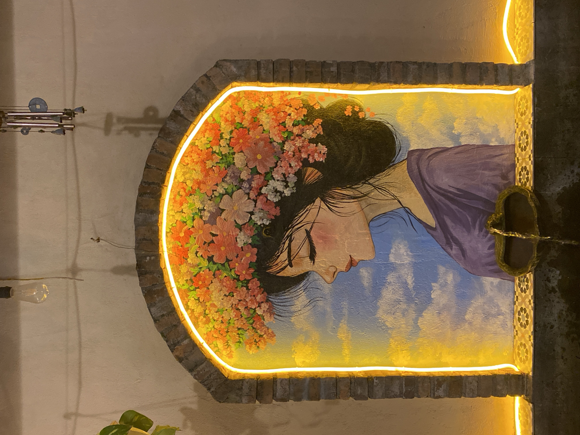 The image depicts a mural of a woman with a floral crown, framed by an arched brick structure. The mural is illuminated by a vibrant yellow neon light that outlines the arch. The woman is painted in profile, with her eyes closed and adorned with a large, colorful flower arrangement on her head. She wears a purple garment. Behind her, the mural features a sky with clouds. Below the mural, there is a small water feature with a decorative tile border. The scene is set against a textured wall. The image is taken from a medium shot, capturing the mural and its immediate surroundings. This mural is located in Nha Trang, Vietnam, specifically at a cafe situated at 26-28 Hùng Vương Street.