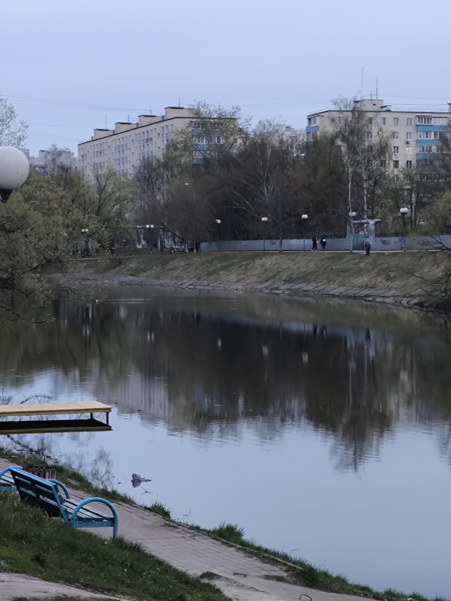 The image presents a serene, overcast scene along a river or canal in Khimki, Russia, likely during early spring given the mixture of budding and bare trees. The sky is a uniform, pale grey, suggesting either dawn, dusk, or a heavily clouded day, casting a soft, diffused light over the landscape.
In the foreground, a paved path lines the near bank, where a distinctive blue park bench with curved metal armrests is situated. A light-colored wooden dock or platform extends from this bank into the calm water, which perfectly reflects the trees and buildings from the opposite side. A small piece of litter is visible floating near the dock.
The midground features the tranquil body of water, leading to the opposite bank. This far bank is also bordered by a paved path, lined with trees varying in foliage density and several streetlights. Three indistinct human figures can be seen walking casually along this path. A notable cross-like structure, possibly a monument or a decorative element, stands out among the trees on this bank. The bank itself has a grassy slope reinforced with concrete sections near the waterline.
In the background, a row of multi-story residential apartment buildings, characteristic of post-Soviet architecture with their repetitive windows and balconies, rise above the trees on the far side. Overhead wires are visible against the pale sky above these buildings. A spherical street lamp fixture is partially visible on the far left edge of the foreground. The overall atmosphere is quiet and tranquil, with muted colors dominating the palette. No text is visible in the image.