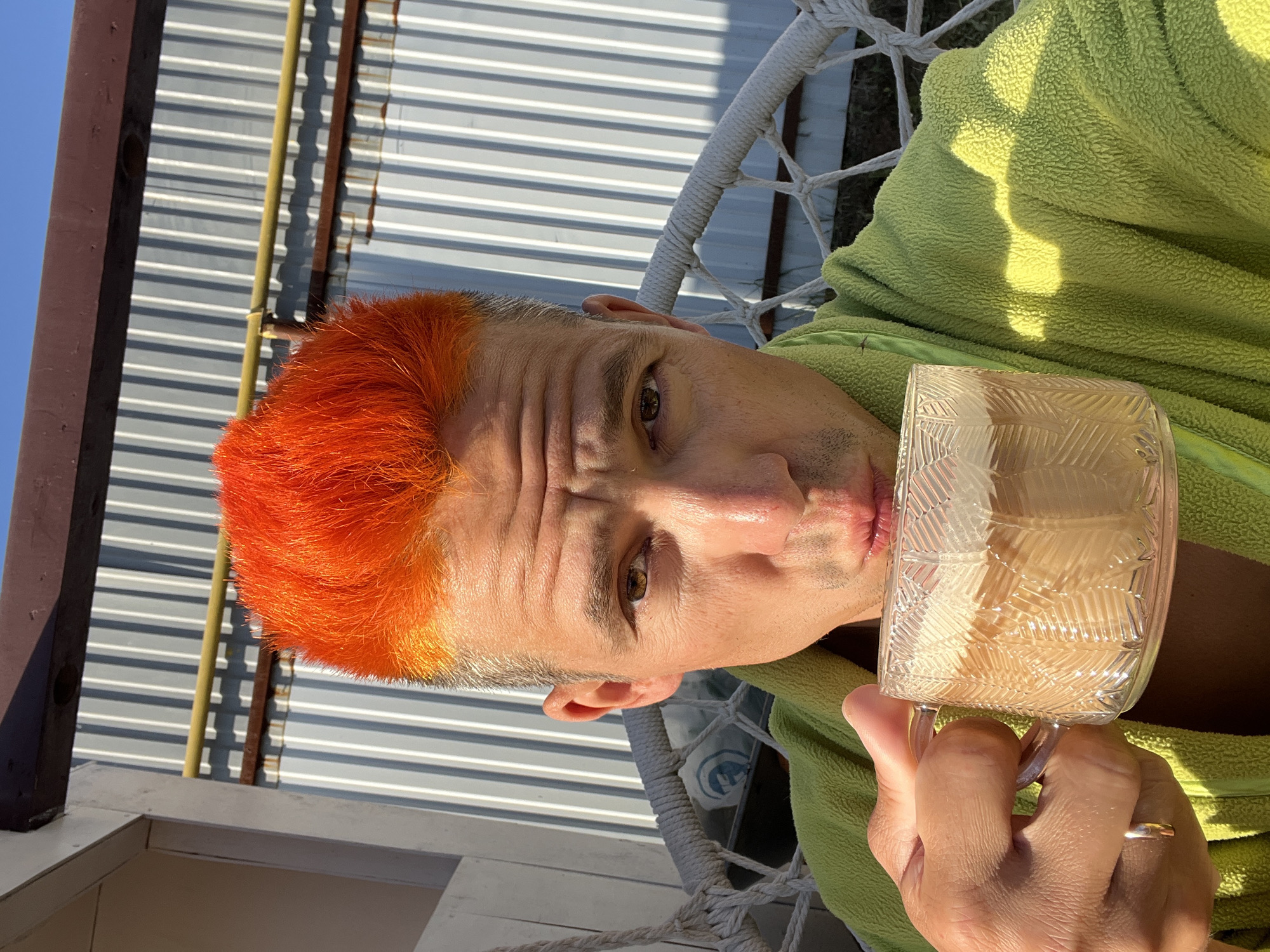 A man with short, orange-dyed hair is reclined in a white wicker chair, possibly a hanging chair. He is wearing a light green terrycloth robe. The man is holding a clear glass mug, possibly containing a light-colored beverage, close to his lips. The background shows corrugated metal siding and what appears to be a blue sky. The setting appears to be outdoors, possibly a balcony or patio. The location is described as Chaikovsky, Russia.