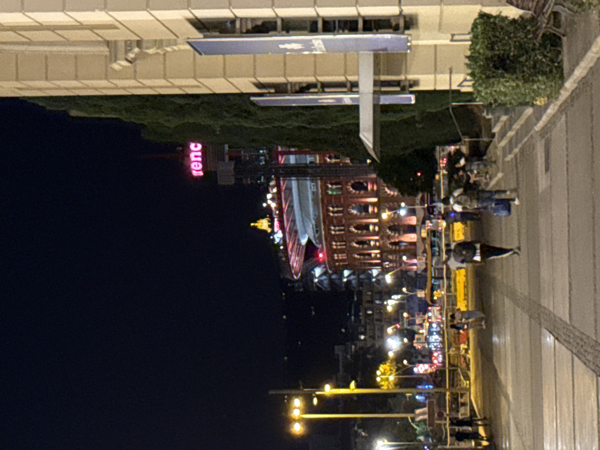 The image captures a nighttime street scene in Barcelona, Spain, likely near the Arenas de Barcelona, a former bullring that has been converted into a shopping center. A large, circular building with illuminated arches and a distinctive domed roof is visible in the mid-ground. The words "renc" in neon pink are lit up on a tall structure to the left of the building. The foreground shows a paved walkway with people walking in various directions. Streetlights cast a warm glow on the scene. To the right, a section of a building with a cream-colored facade and vertical blue banners is prominent. The overall atmosphere is that of a bustling urban environment at night.