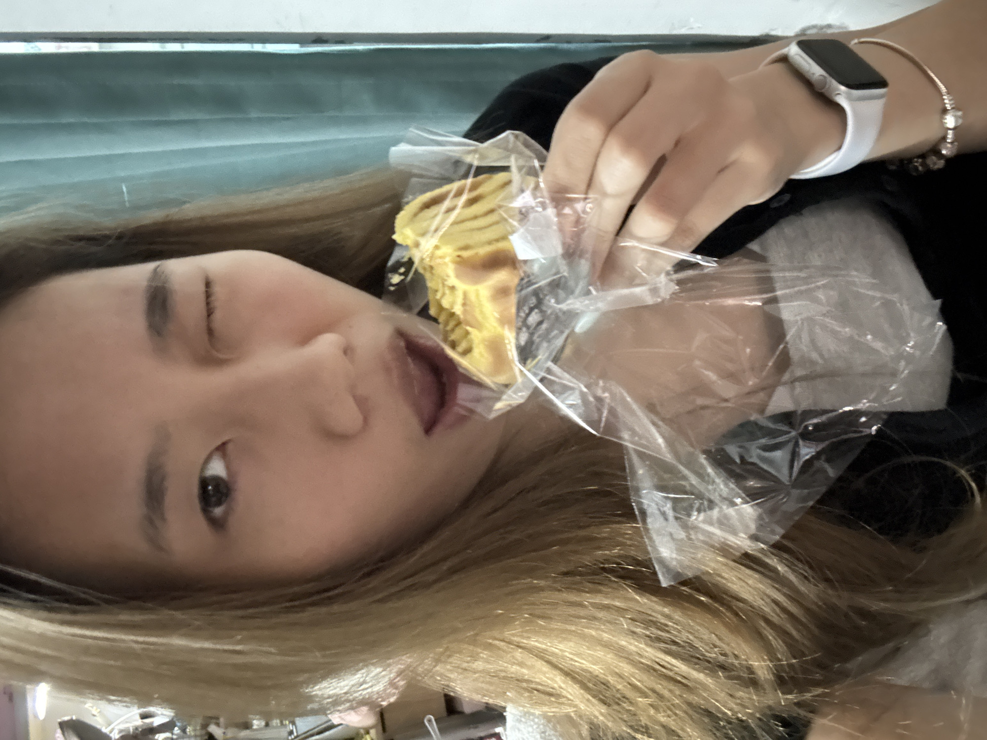 A light-skinned woman with blonde hair is holding a pastry in a plastic wrapper, poised to eat it. She is winking with her left eye. She wears a smartwatch and a beaded bracelet on her left wrist. The background features a light blue curtain or wall. The location in the city of Nan'an District, China is not discernable from the given image.