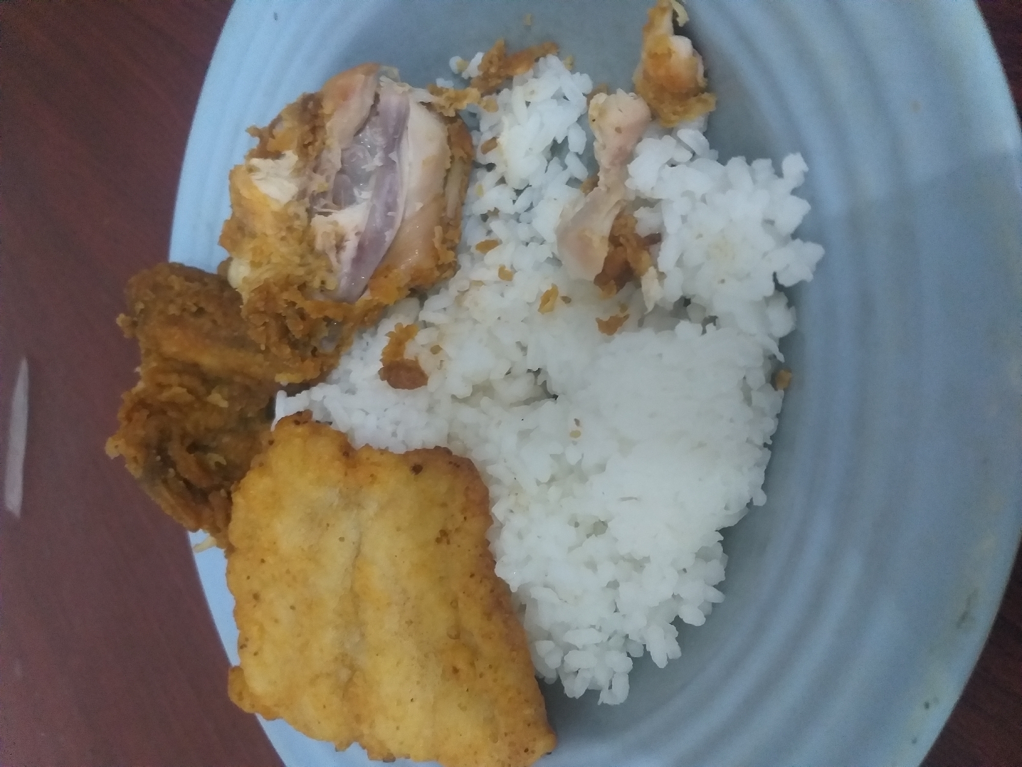A plate of food containing white rice, a piece of fried fish, and fried chicken is presented on a blue plate. The plate is placed on a wooden table. The scene is likely in a home setting in Air Jamban, Indonesia.