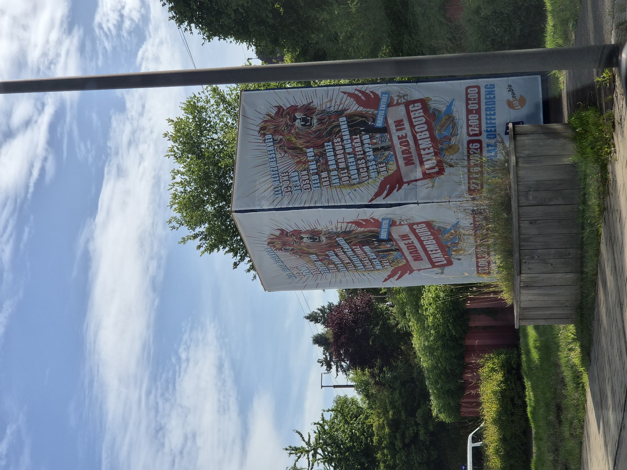 A tall, white, rectangular advertisement with red lettering and images of lions is displayed outdoors. The advertisement is located in Differdange, Luxembourg. The ad displays the text "MADE IN LUXEMBOURG" prominently in red. Below this, it reads "27.06.2026 17:00-01:00" and "PLATZ DEIFFRDENG". A logo for "Eldorado Radio" is visible on the bottom right of the advertisement.
The advertisement is situated next to a grey pole and surrounded by greenery, including trees and bushes. In the background, there are more trees and a blue sky with white clouds. A wooden planter box filled with grass and flowers is in the foreground. A white vehicle is partially visible to the left of the advertisement. The ground in front of the planter is paved with grey bricks.