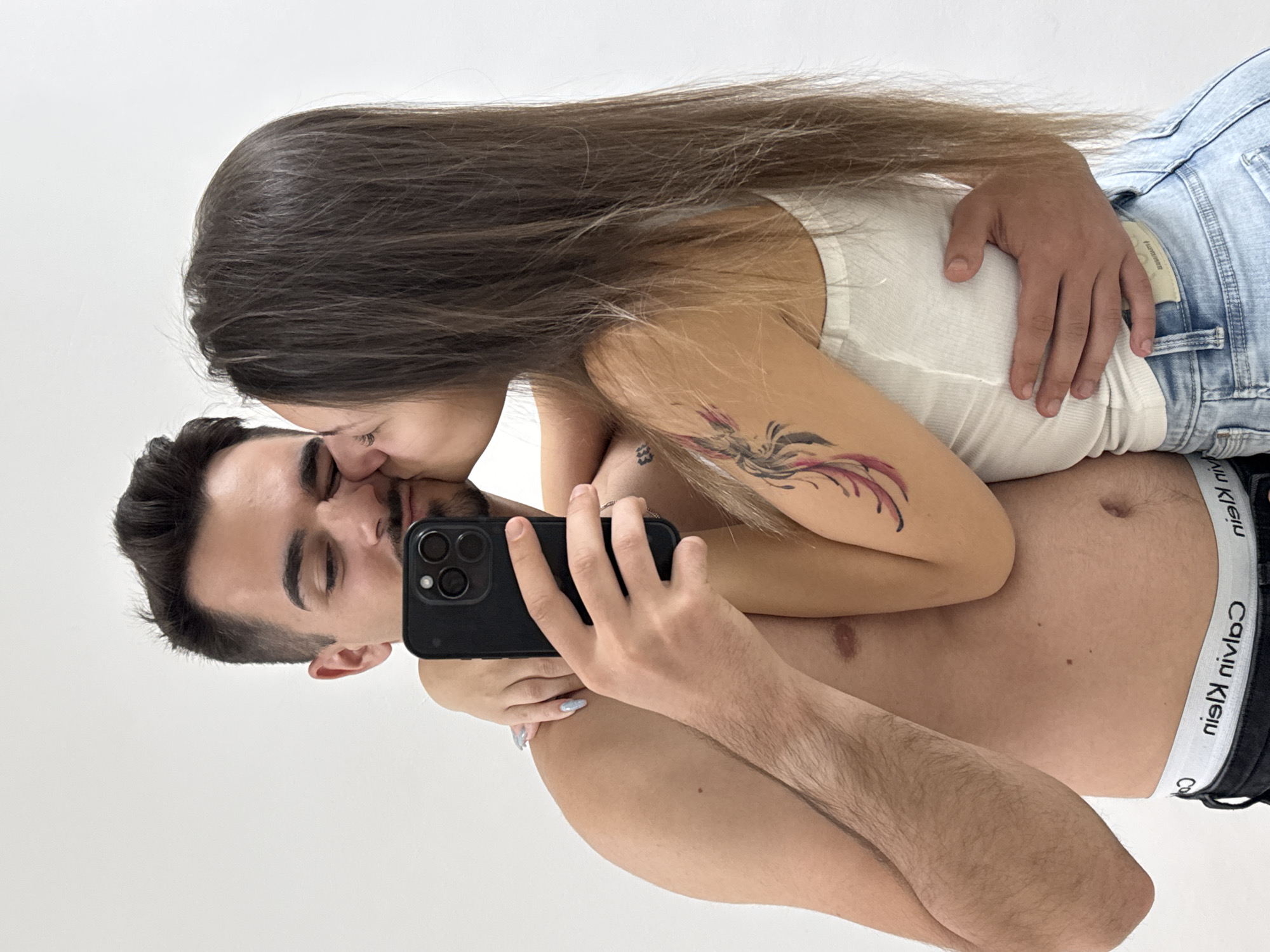 The scene takes place in an indoor setting in Sofia, Bulgaria, characterized by a plain white background. The image is a selfie of two individuals, a male and a female, in a close embrace. The male, positioned on the left, is shirtless with dark hair and stubble. He wears black Calvin Klein underwear with the brand name "nie⋊ nivlɒƆ" visibly reversed on the waistband, indicating a selfie or mirror image. He holds a black smartphone with a prominent triple-lens camera module in his right hand, angled to capture the shot. His left arm is wrapped around the female's waist. The female, on the right, has long, straight dark brown hair. She wears a white ribbed tank top and light blue denim jeans. A large, multi-colored tattoo resembling a phoenix is visible on her upper left arm, and a smaller, indistinct tattoo is present on her inner left forearm. Her right arm is wrapped around the male's neck/shoulder, and her left hand is placed on his lower back. She is kissing the male on his right cheek. The female's visible fingernails are painted with light blue polish. The lighting is bright and even, without discernible harsh shadows.