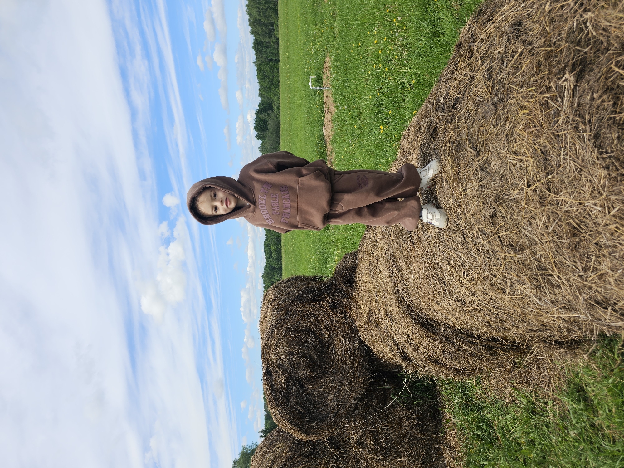 The image shows a young child in a brown hooded sweatshirt and matching pants standing on a hay bale in a grassy field. The child is wearing white shoes. Behind the child is a green field that extends into the distance. The sky is blue with white clouds. The scene is likely located in a rural area, specifically Borodino, Russia, as specified. There are other hay bales visible at the bottom.
