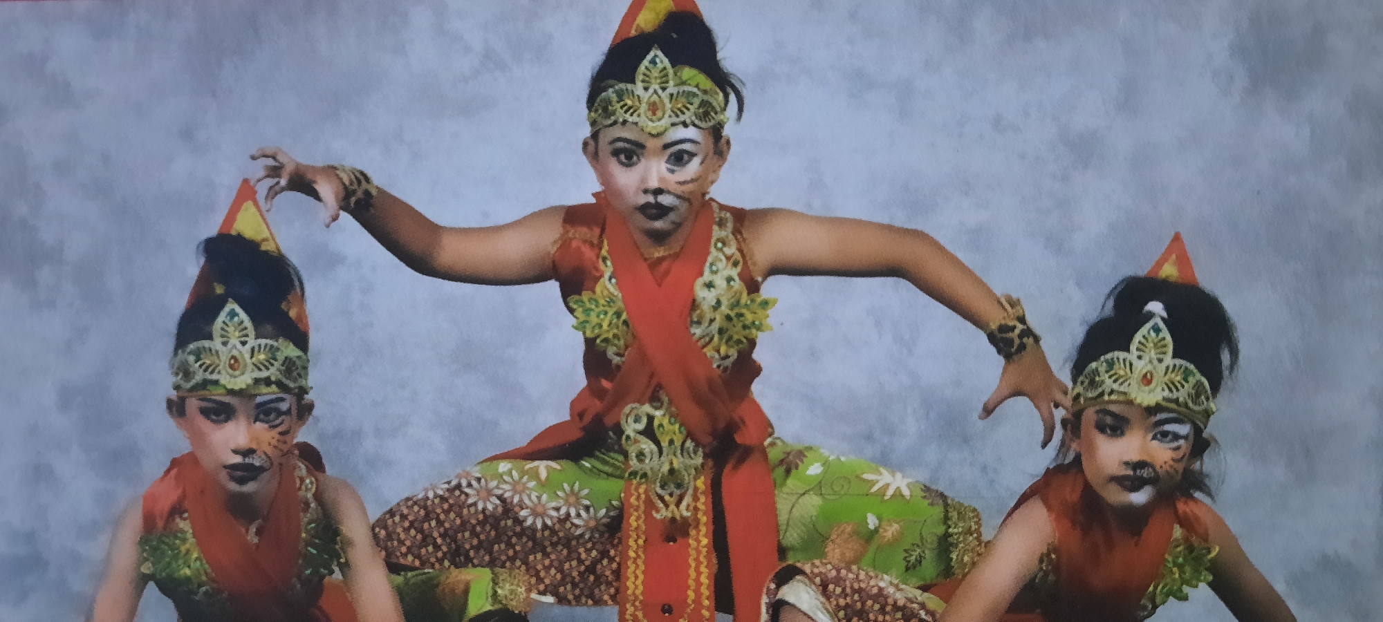 This image shows three children dressed in traditional Indonesian dance costumes. The child in the center is larger and positioned in the foreground, while two smaller children are positioned on either side, slightly behind. All three children have their faces painted with dark eye makeup and designs resembling a tiger's face, with black whiskers and markings. They are all wearing conical headdresses with decorative elements and ornate bodices. The central figure wears a bright orange sash over a green and brown patterned skirt. The children on the sides wear similar costumes with variations in color and pattern. Their poses suggest they are in the midst of a performance. The background is a mottled gray, suggesting a studio setting or backdrop. The children appear to be participating in a cultural event, possibly a dance recital or performance, in the city of Ploso, Indonesia.