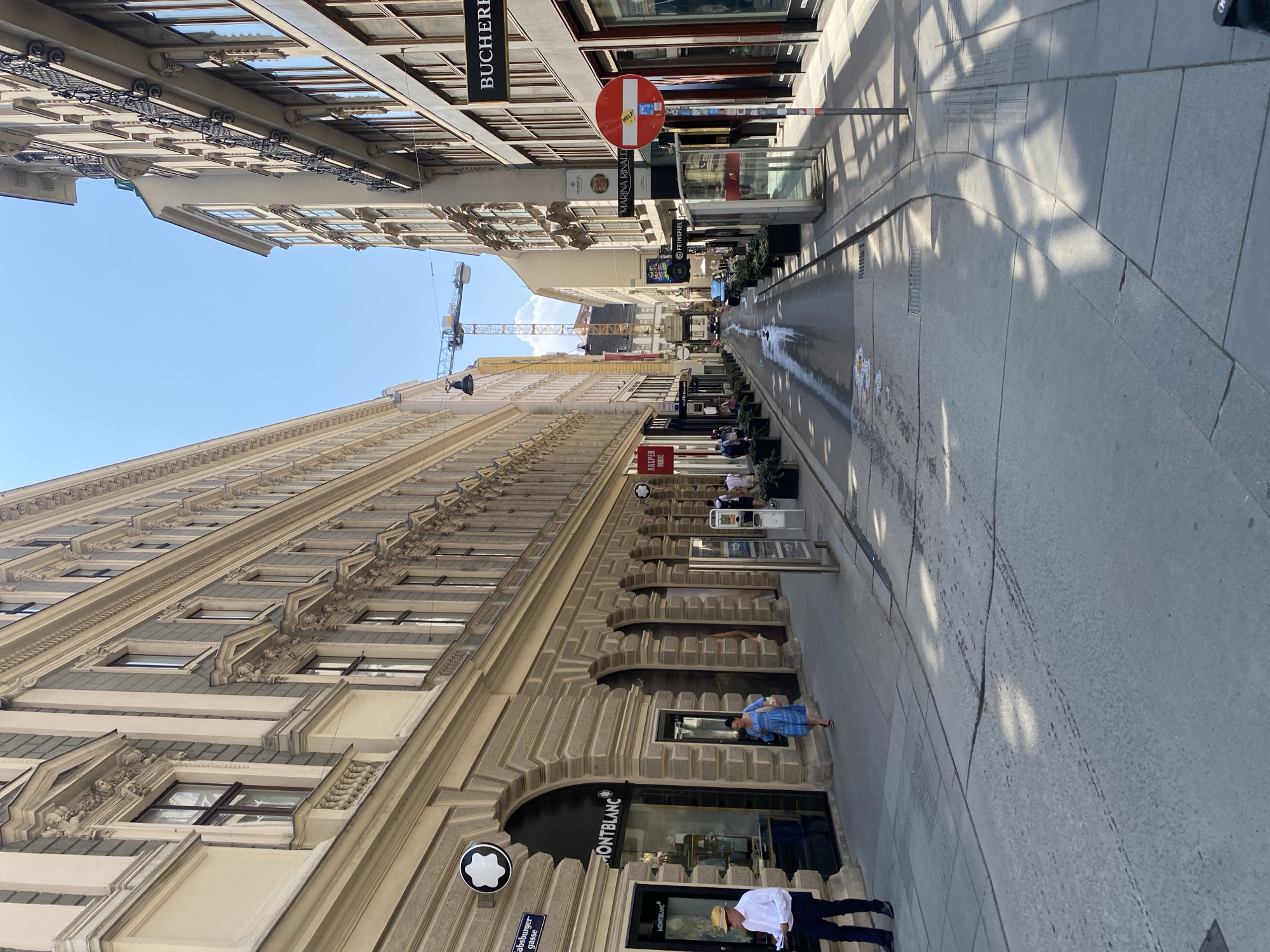 The image captures a street scene in Vienna, Austria, with a view down a narrow street lined with ornate buildings on both sides. The architecture suggests a historic European city, possibly with shops at street level and residences or offices above. Sunlight casts sharp shadows on the cobblestone street, indicating it is a bright, clear day.
On the left side of the street, a Montblanc store is visible, indicated by its prominent signage and a circular logo above the entrance. A man in a white shirt and dark pants stands near the store entrance, holding some papers. Further down, a woman in a blue dress walks along the sidewalk.
The right side of the street also features shops, with signage for "BUCHERER" being clearly visible. A red and white circular traffic sign is posted on the right sidewalk.
In the distance, a construction crane is visible, suggesting ongoing development or renovation in the area. The street itself appears to be a pedestrian zone or a street with limited vehicle traffic, as indicated by the presence of planters and the overall ambiance. The ground is a mix of pavement and what appears to be cobblestones or large stone slabs. There are some wet patches on the street, possibly from recent cleaning or a spilled liquid. The sky is a clear, vibrant blue.