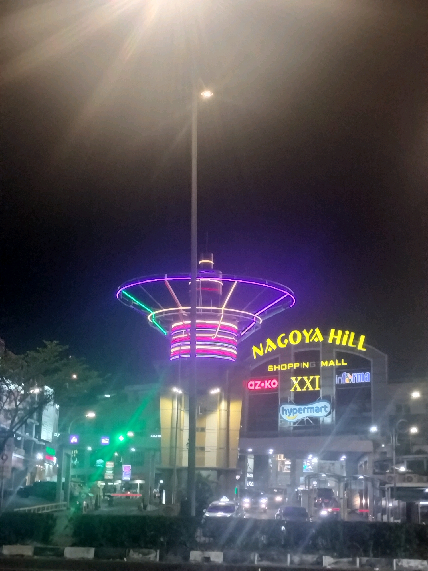 The image depicts the Nagoya Hill Shopping Mall in Batam, Indonesia, at night. A tall street lamp with a bright light emanates from the top of the frame, casting a lens flare downwards. In the foreground, a multi-tiered, brightly lit structure with neon accents of purple, green, and pink stands prominently, resembling a modern art installation or decorative element. The Nagoya Hill Shopping Mall sign is visible in large yellow lettering against the building's facade, with smaller signs below indicating "SHOPPING MALL," "az-ko," "XXI," and "informa," as well as a "hypermart" logo. The area in front of the mall features traffic lights, with cars and headlights visible on a road. Trees line the street, and the overall scene is illuminated by the artificial lights of the mall and street lamps, creating a vibrant urban nightscape.