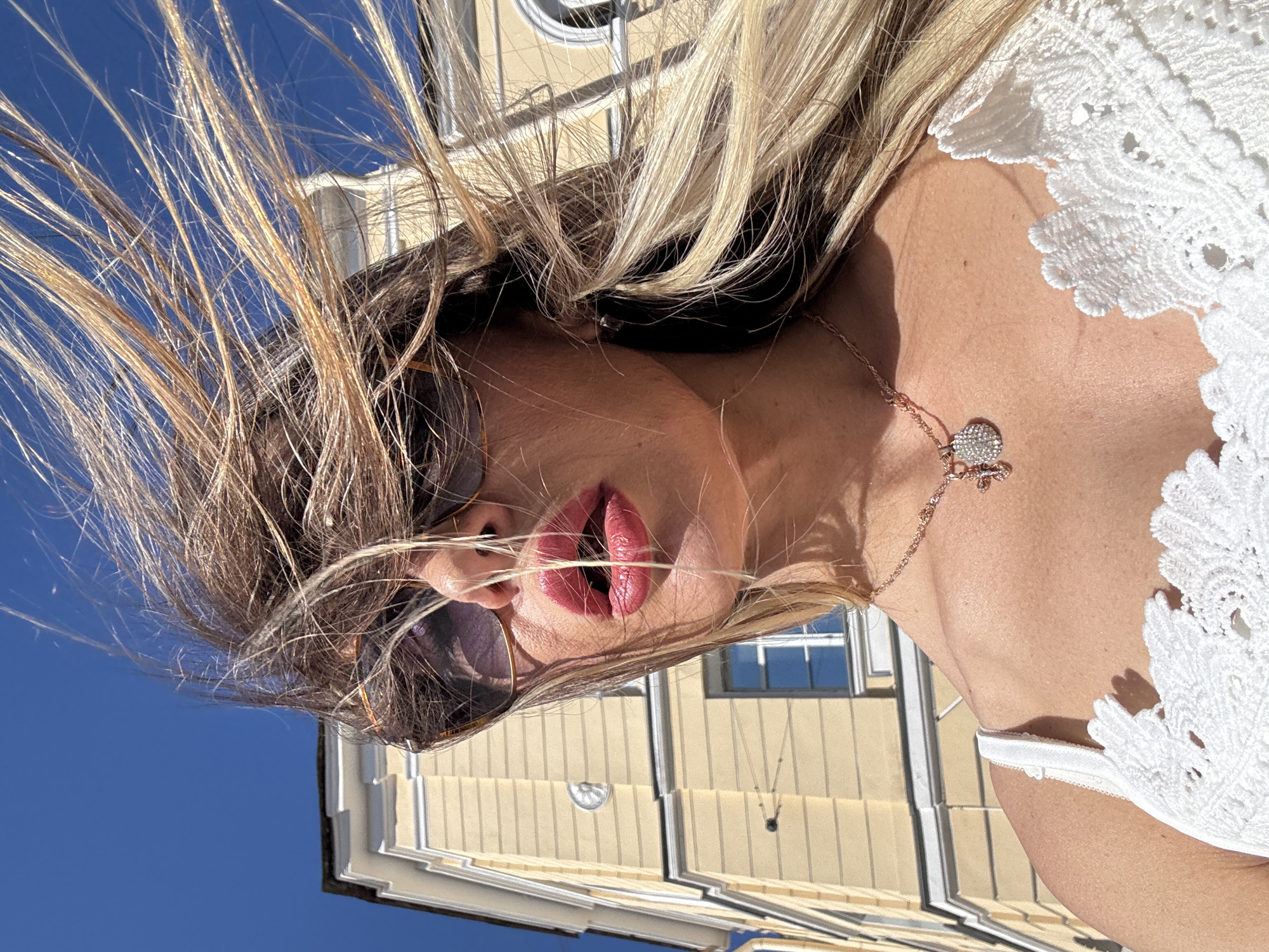 A close-up, low-angle shot shows the face of a woman with blonde hair blowing in the wind. Her lips are painted with a glossy pink lipstick, and she is wearing sunglasses. A rose gold necklace with a heart-shaped pendant rests on her chest, which is adorned with white lace detailing. The background features a light yellow building with a blue sky above. The sunlight suggests it is daytime, likely a clear and warm day. The scene is likely outdoors, given the wind and bright sunlight. The woman appears to be posing for a selfie. The architectural style of the building may suggest a European location, possibly Saint Petersburg, Russia, given the context provided. No visible text is present in the image.