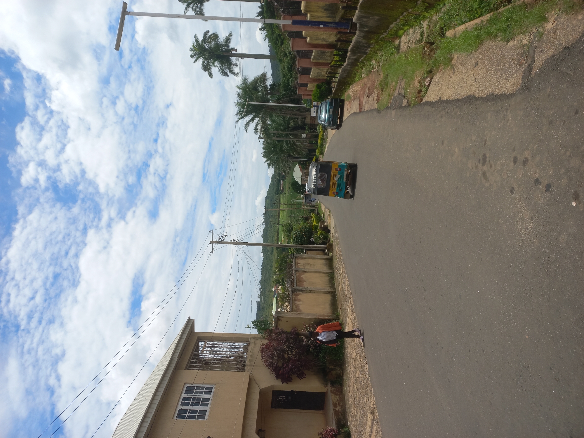 A street scene in Jos, Nigeria, under a partly cloudy sky. A two-story tan building with white-framed windows is on the left, with a black door and a red-leafed bush in front. A woman wearing an orange jacket, white shirt, and dark pants walks on the sidewalk to the right of the building. Further down the paved road, a yellow and blue auto-rickshaw is in motion, followed by a dark green sedan. The road is bordered by a beige concrete wall on the right, overgrown with grass and moss. Palm trees and other lush green vegetation line the background, with rolling hills visible in the distance. Electrical poles and wires are present throughout the scene.